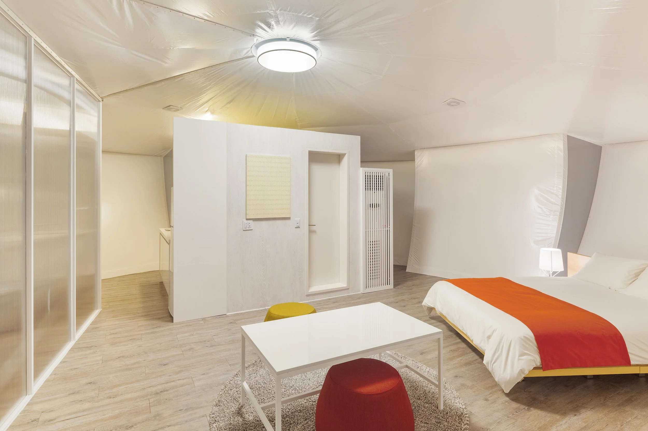 Minimalist bedroom with white walls, a bed with white linens and an orange throw, a small white table, and colorful stools, with unique curved ceiling and modern lighting.