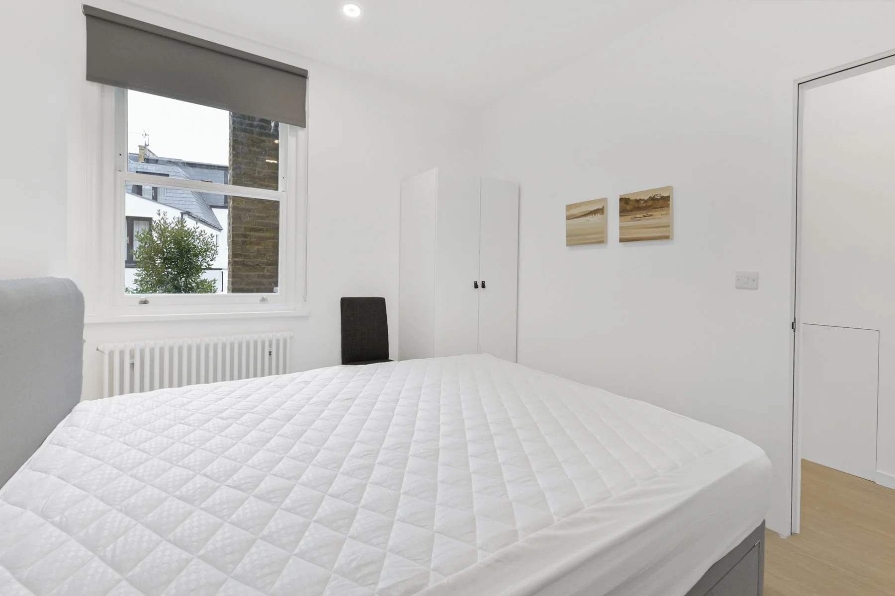 A minimalistic bedroom with a white quilted bed, black and gray chairs, a white wardrobe, a window with a view of a neighboring building and green tree, and beige wooden flooring.