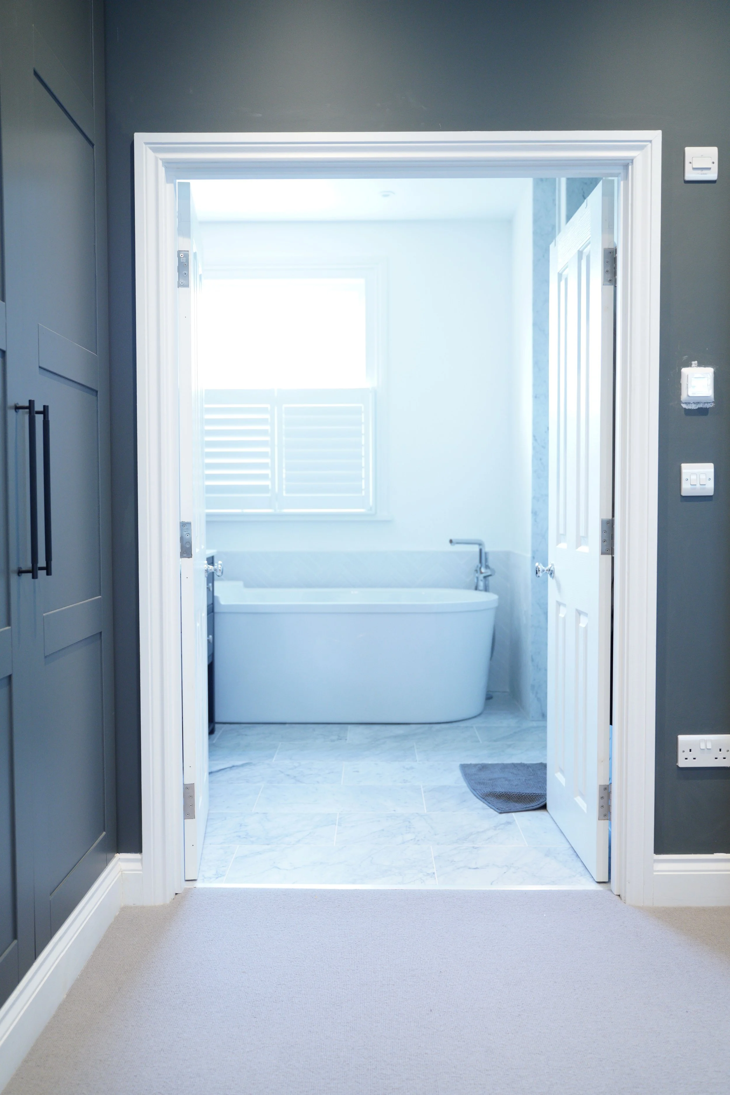 View into a bright bathroom with a white bathtub, window with shutters, and marble flooring, seen through an open door from an adjacent room with dark walls.