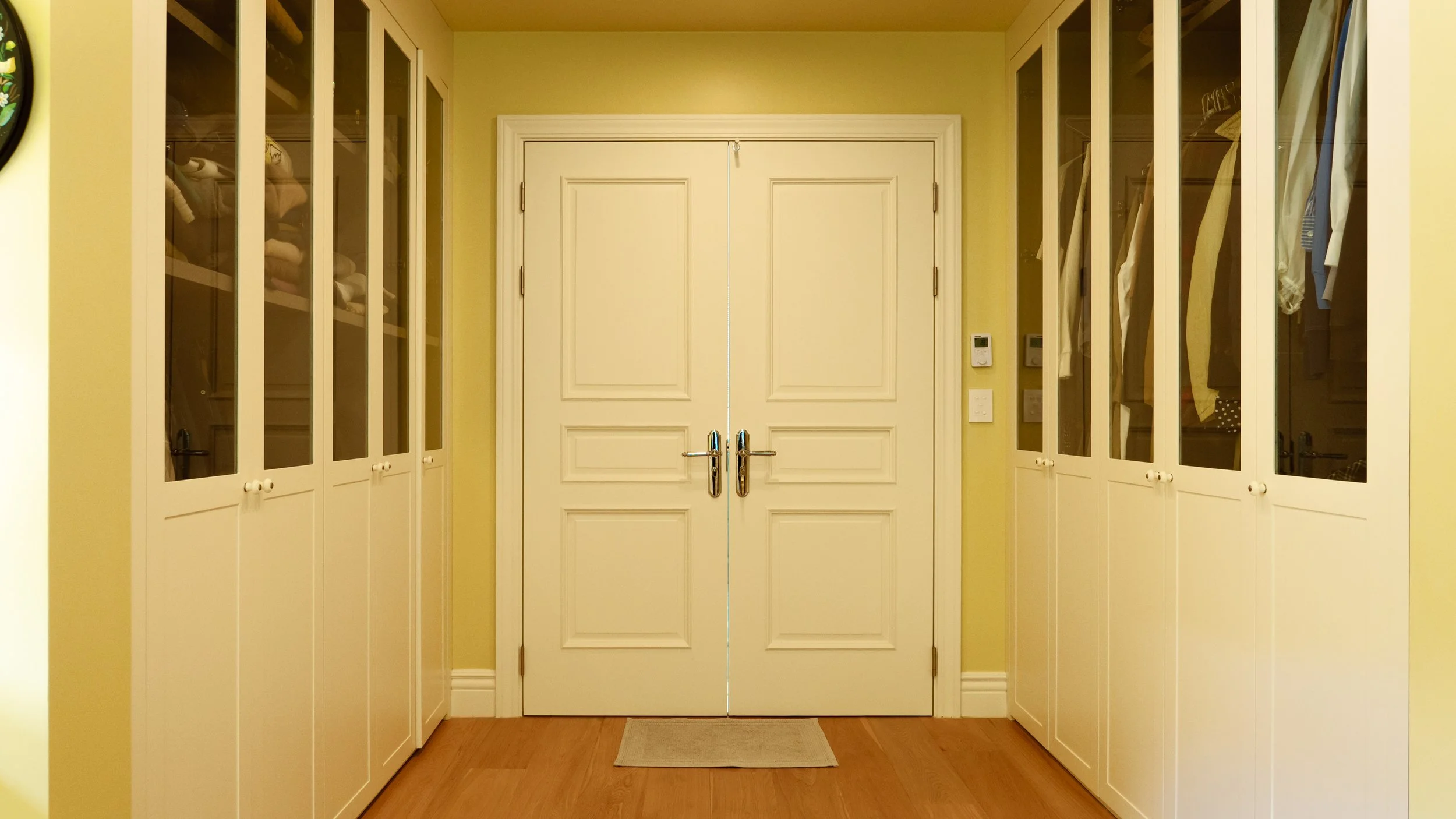 Entryway with double white doors and built-in white cabinets with glass doors on both sides, containing clothes and household items, with a small beige rug on a hardwood floor.