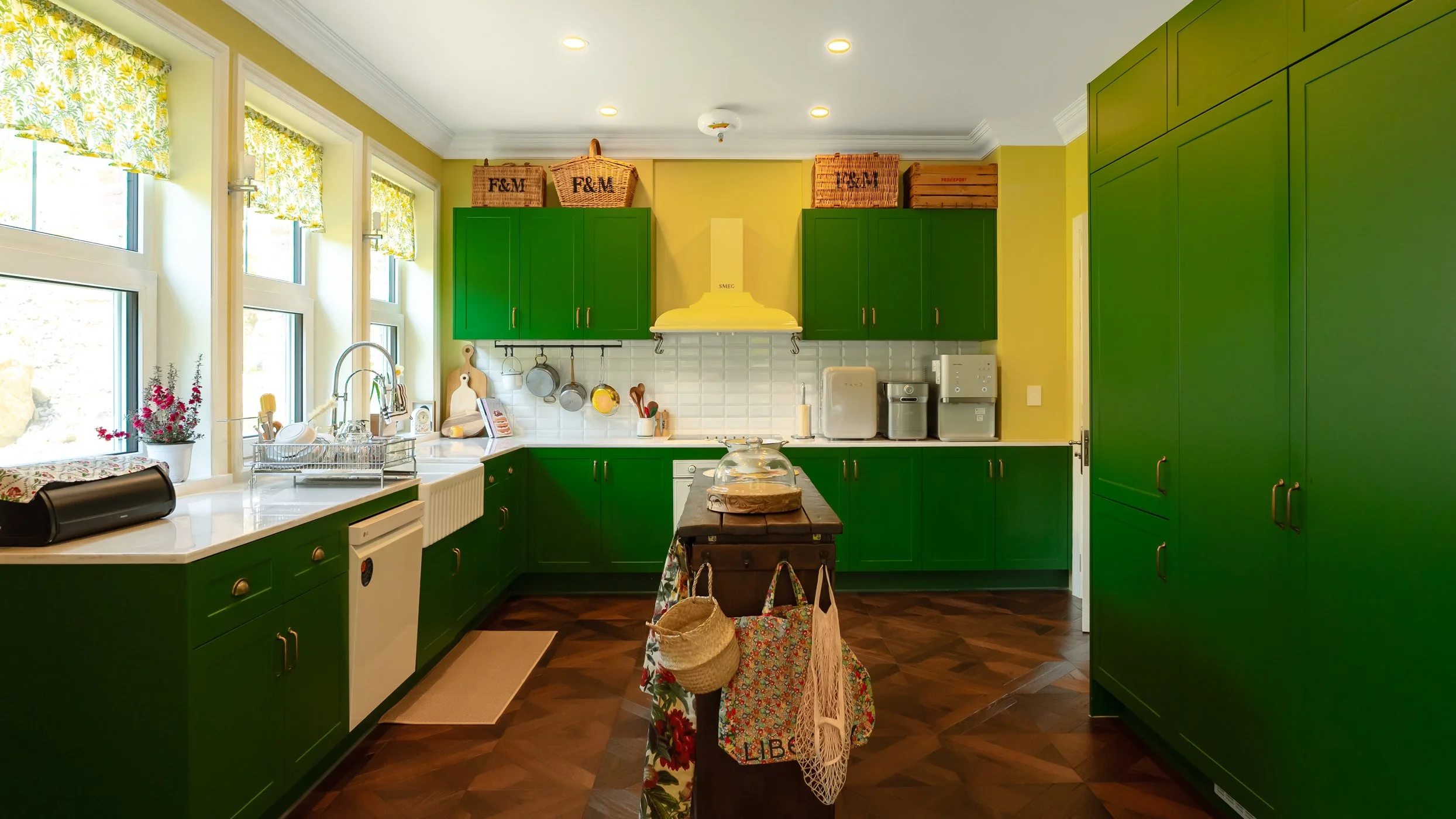 Bright kitchen with yellow walls, green cabinets, a large window with floral curtains, and dark wood flooring.