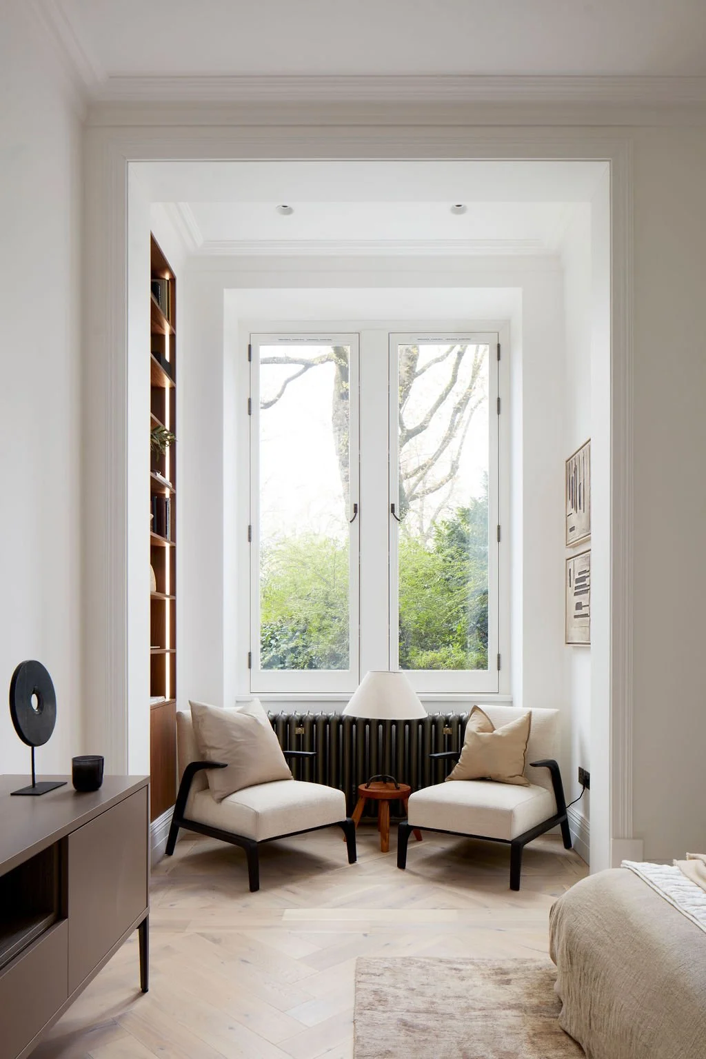 A cozy sitting area by a large window with greenery outside. Two white armchairs with beige cushions are positioned on either side of a small wooden side table with a white lampshade. A black radiator is underneath the window, and a dark wood bookshelf is on the left wall. The room has white walls, light wood flooring, and minimalistic decor.