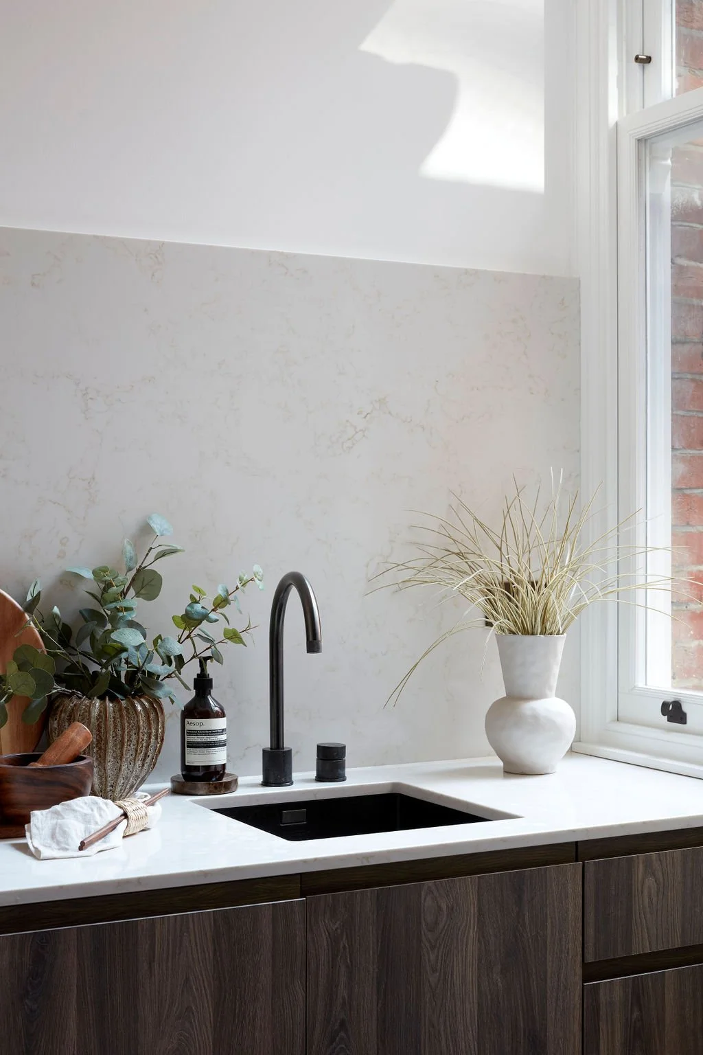 A kitchen countertop with a black sink, a black faucet, and a potted plant with dried grass on a windowsill. There are also a smaller plant in a brown vase, soap dispenser, and a wooden bowl with utensils.