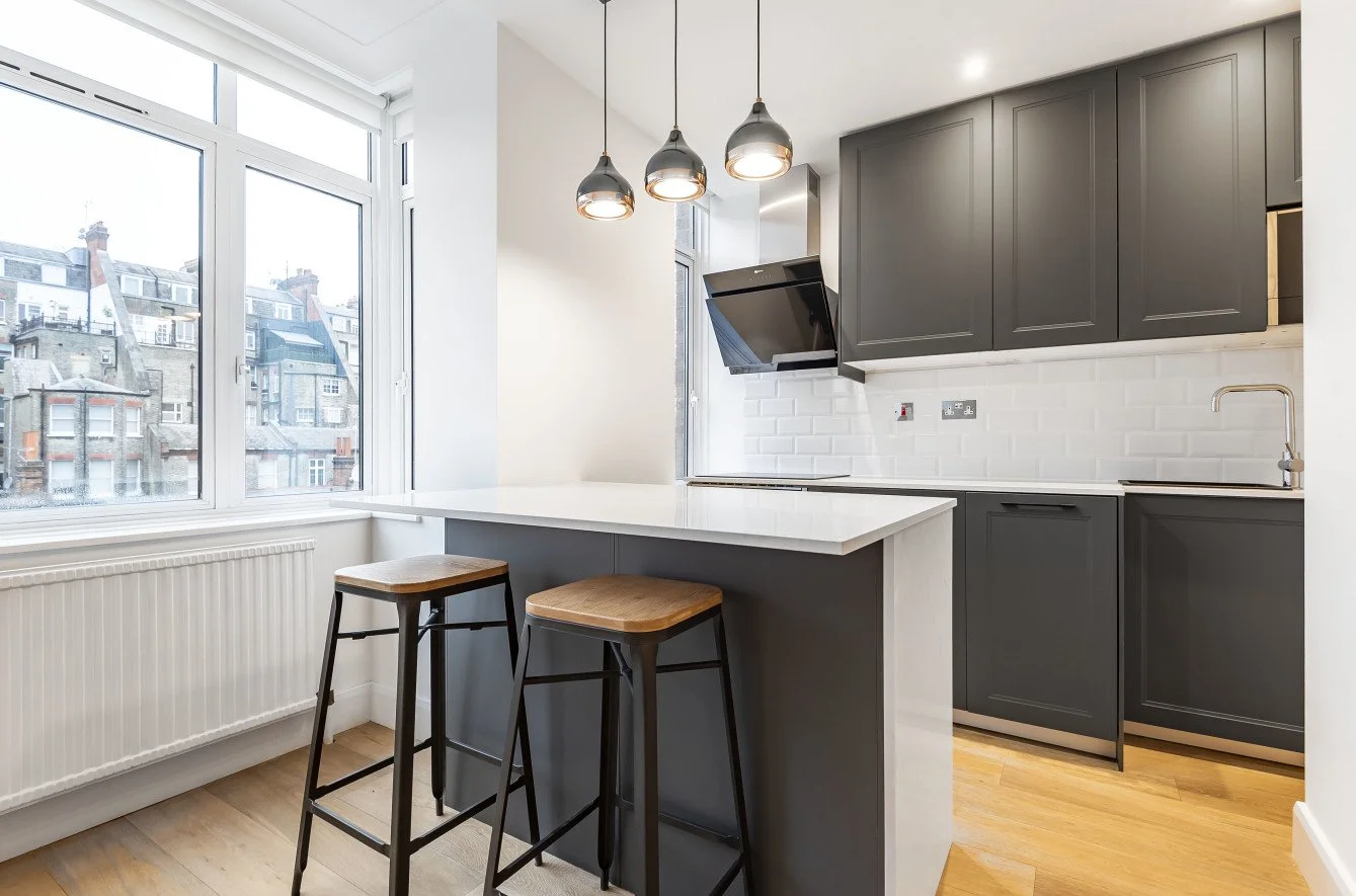 Modern kitchen with dark gray cabinets, white countertops, brick backsplash, wooden bar stools, large window with city view, and hanging pendant lights.