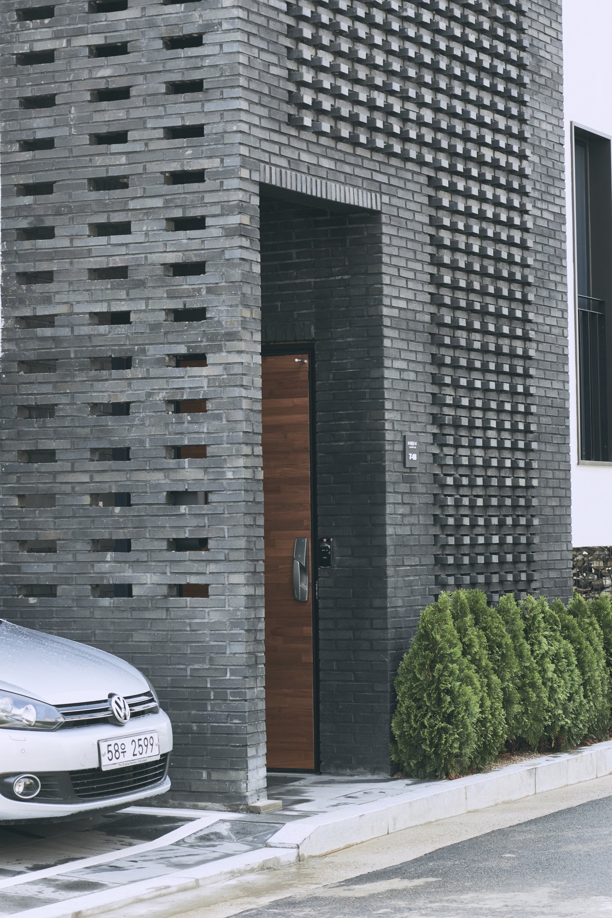 Modern gray brick house with a wooden front door, a parked silver Volkswagen car, and green shrubs in front.
