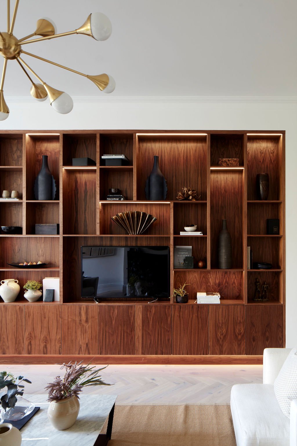 Living room with a large wooden shelving unit decorated with vases, sculptures, and books, and a TV in the center. A white sofa is partially visible in the foreground, and a coffee table with a vase and plants is in front.