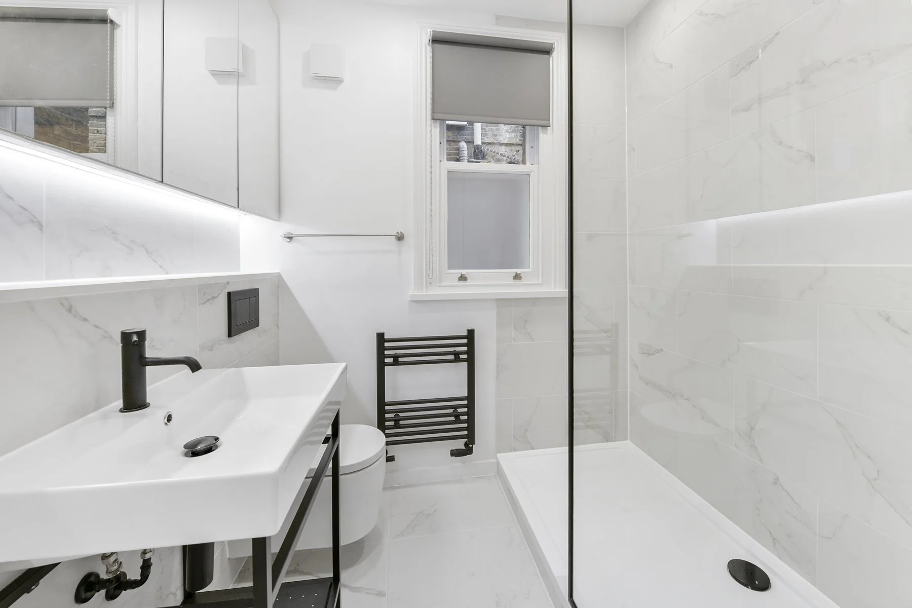Minimalist bathroom with white marble tiles, black fixtures, a small sink, window with gray blinds, and a shower area with a glass partition.
