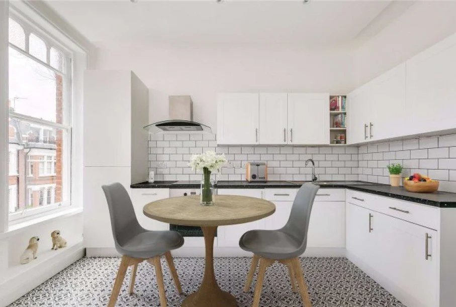 Bright kitchen with white cabinets, black countertop, white subway tile backsplash, round wooden table with two gray chairs, window with decorative figurines, vase of flowers on table, fruit bowl on counter.