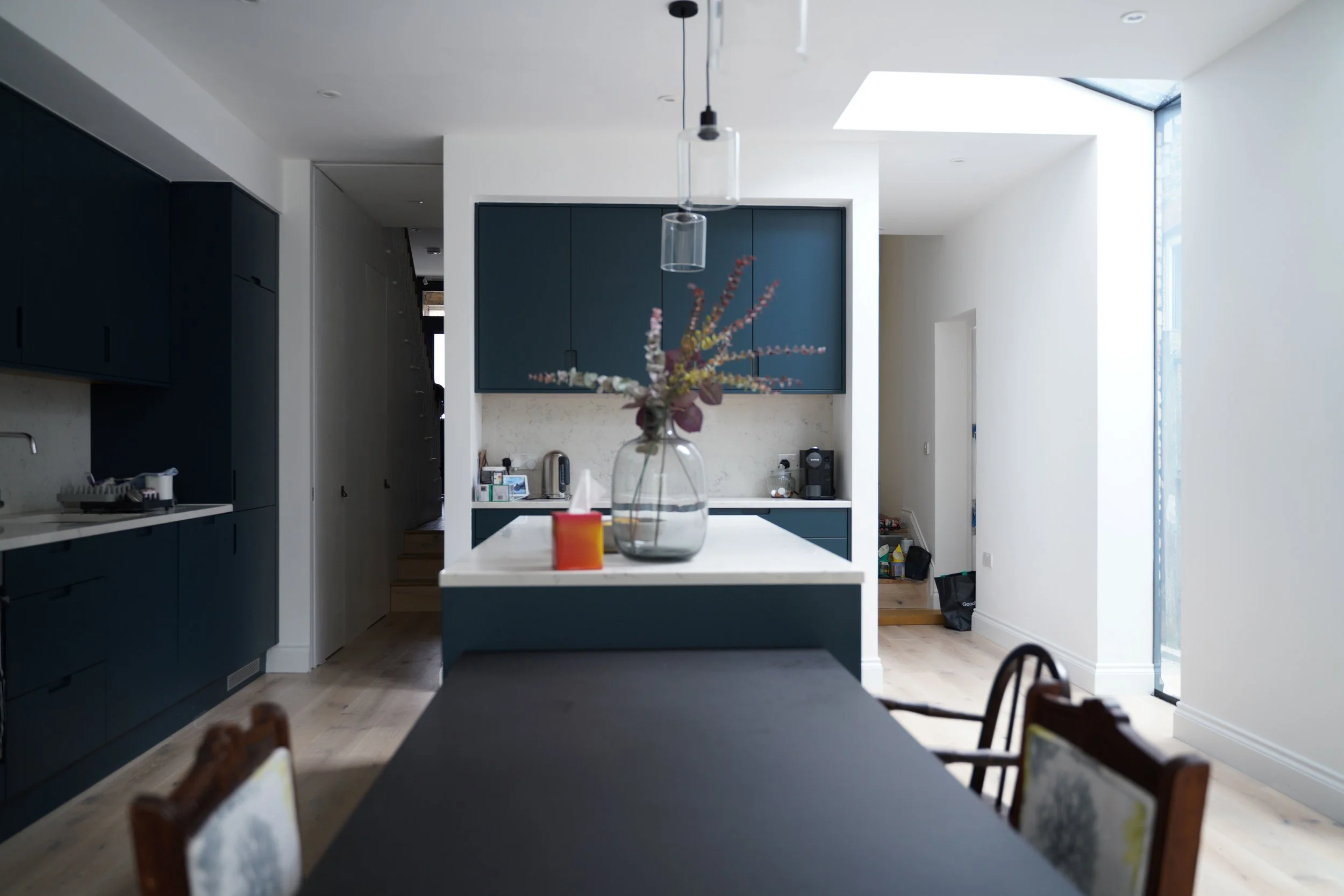 Modern kitchen with dark blue cabinets, white island with a glass vase and flowers, dining table with chairs, and bright natural light from large windows.