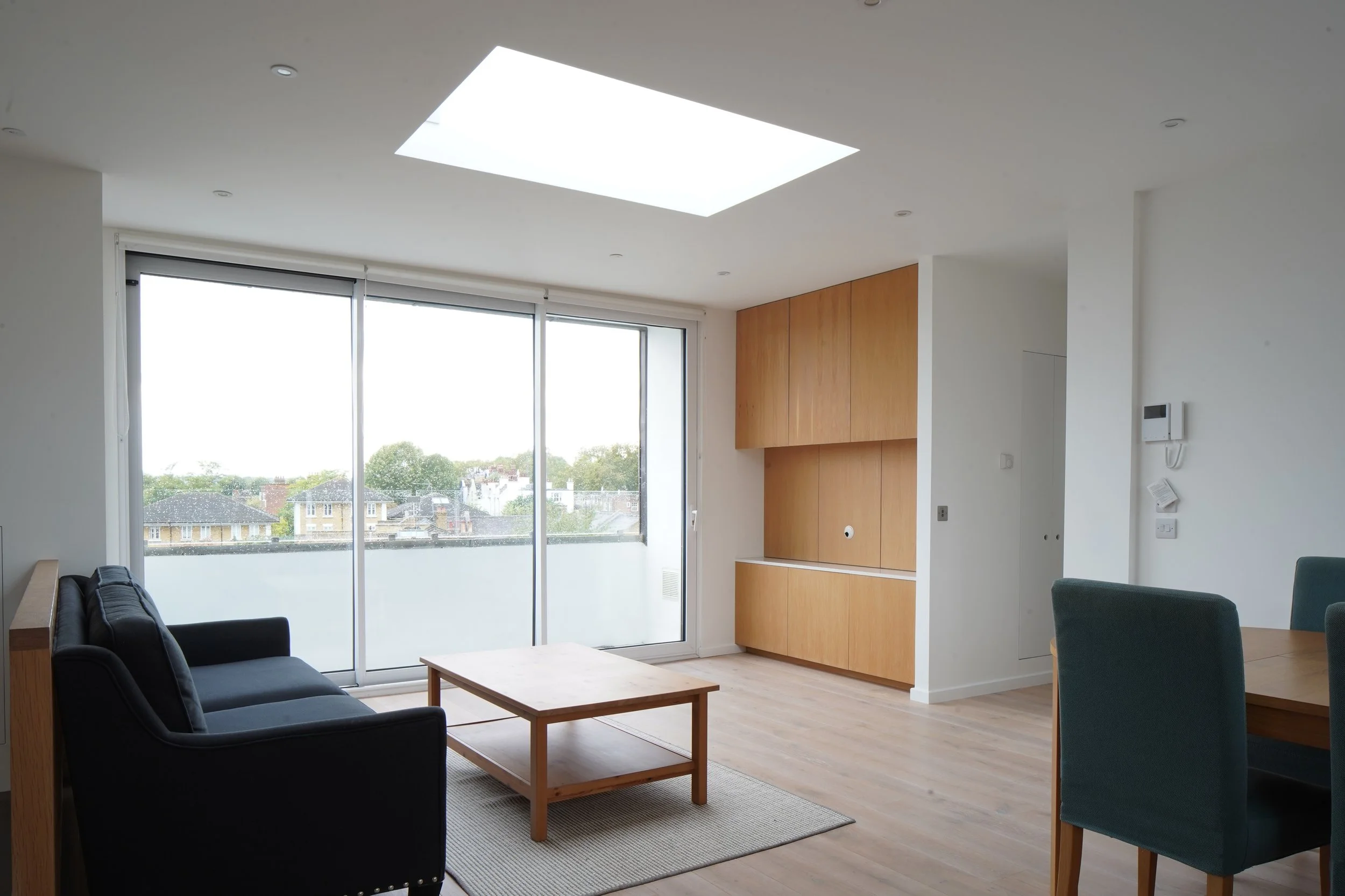 Modern living room with large sliding glass door, wooden cabinets, black sofa, wooden coffee table, and green dining chairs, overlooking a cityscape.