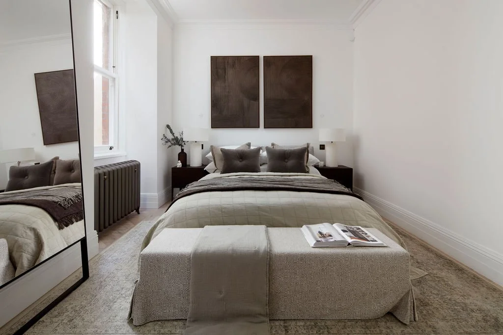 A neatly made bed with beige and gray bedding, two gray pillows, and a beige bench at the foot of the bed in a minimalist bedroom with white walls, a large mirror, and black nightstands with white lamps.
