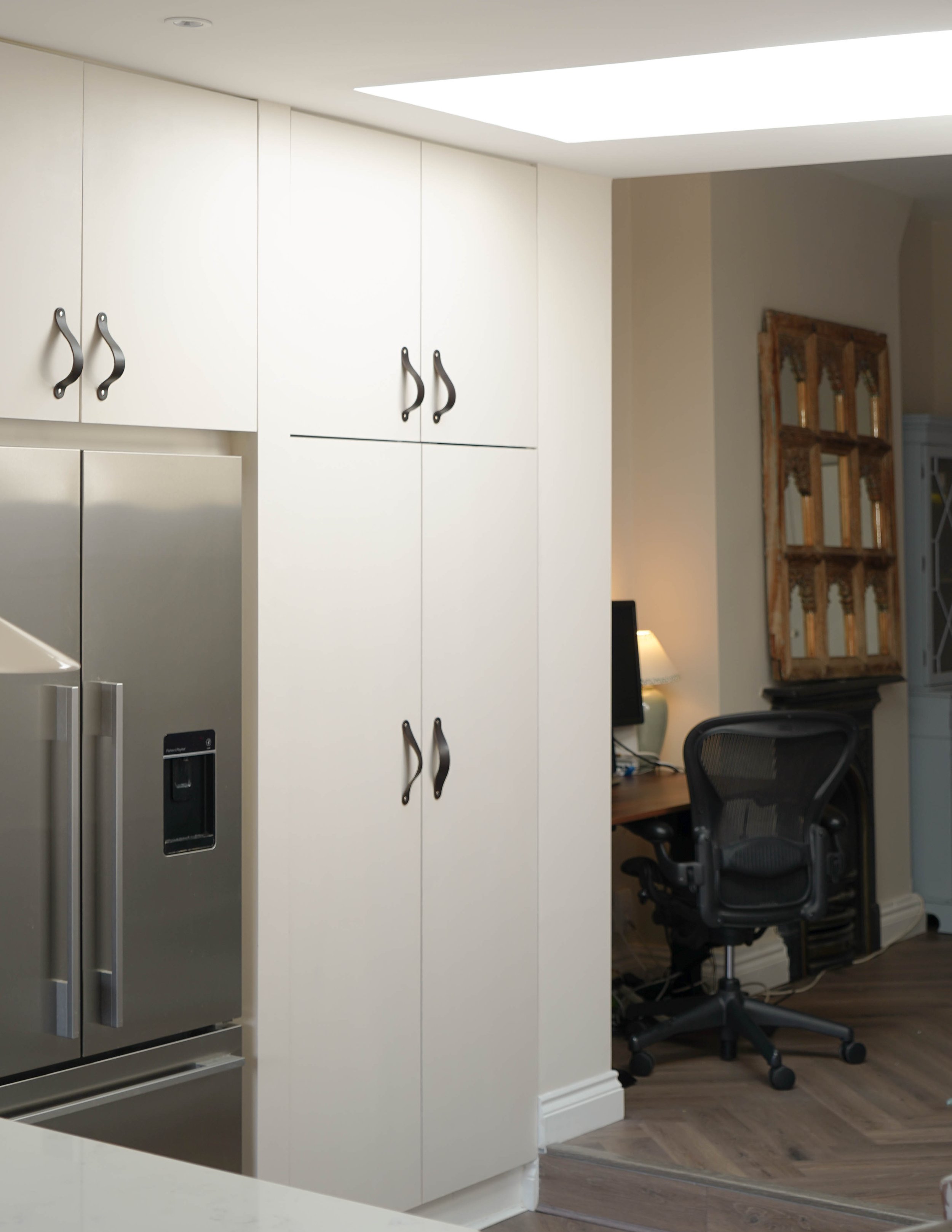 View of a kitchen corner with white cabinets, a stainless steel refrigerator, and a home office setup with a desk, computer, and chair in the background.