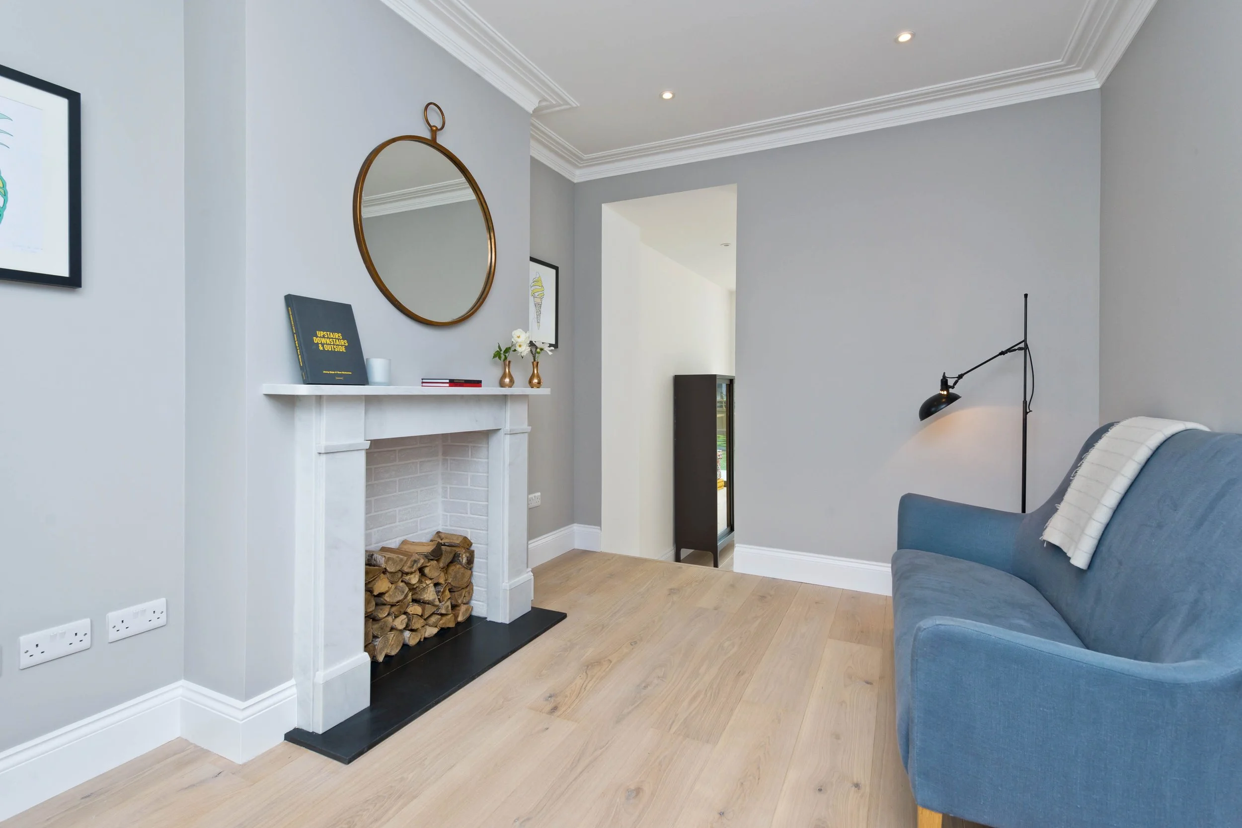 Living room with grey walls, a blue sofa, a black floor lamp, art on the walls, and a white fireplace with stacked firewood inside.