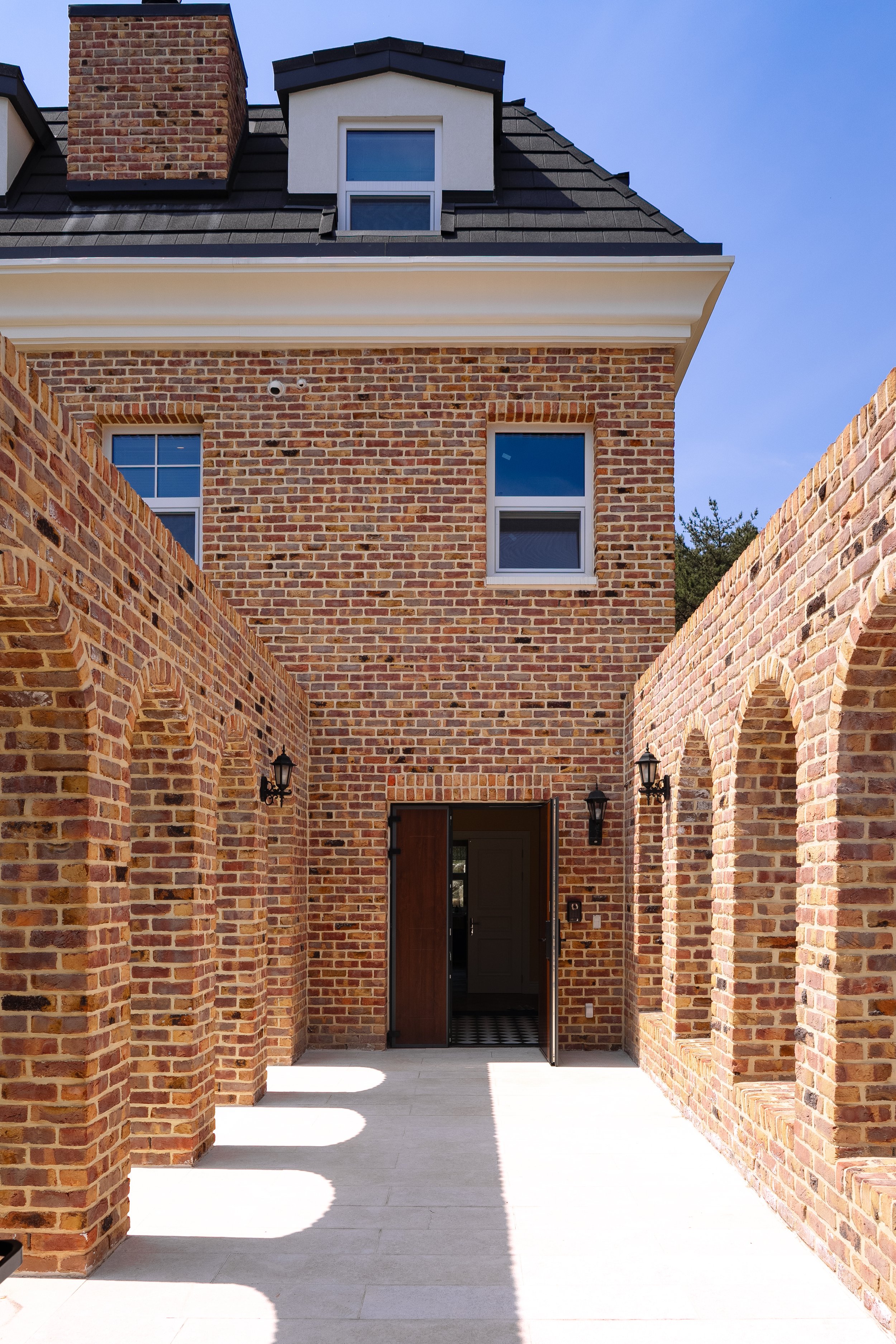 Brick residential building with arched windows on right and a door in the center, outdoor corridor with shadow patterns, and a two-story window, under a clear blue sky.