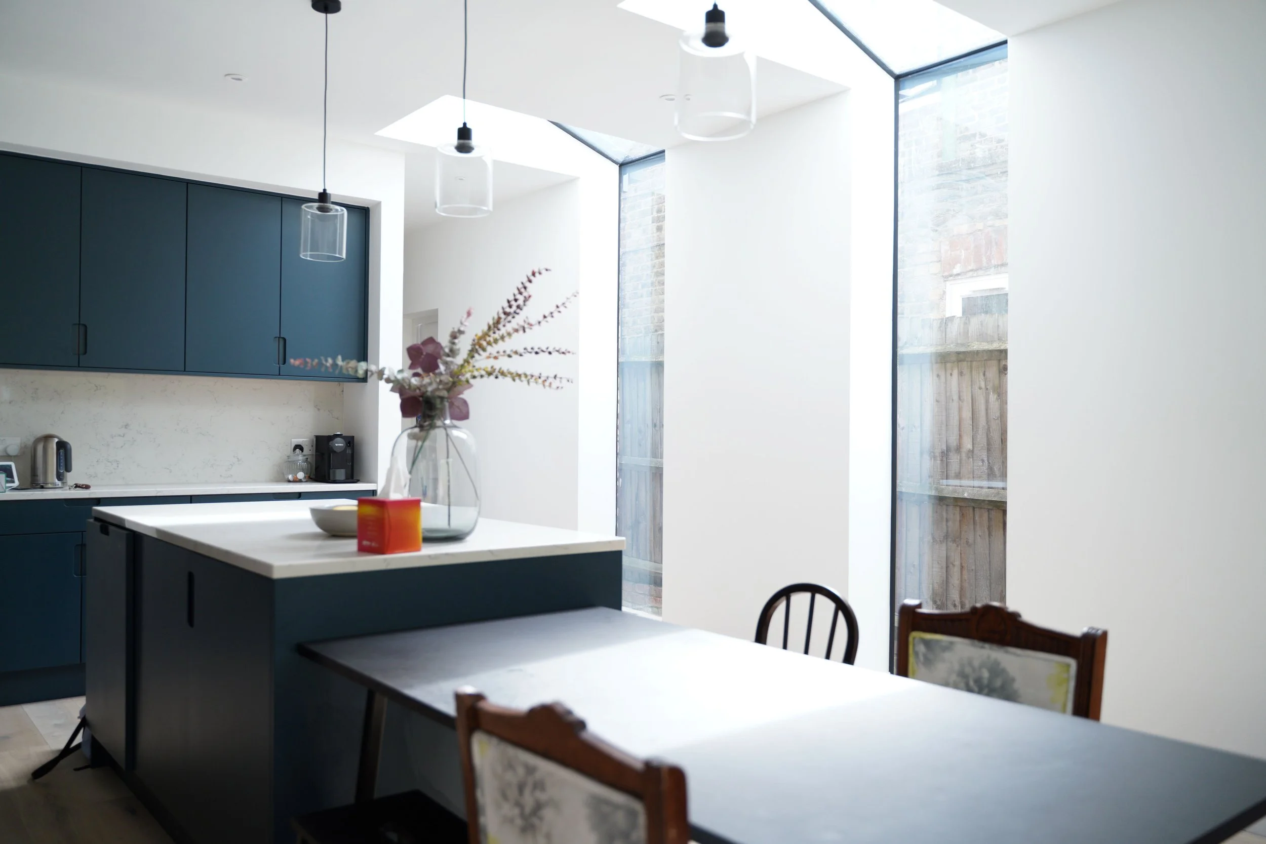 A modern kitchen with white walls, blue cabinets, a white island with a glass vase and floral arrangement, and a dining table with vintage chairs. Natural light from large windows illuminates the space.