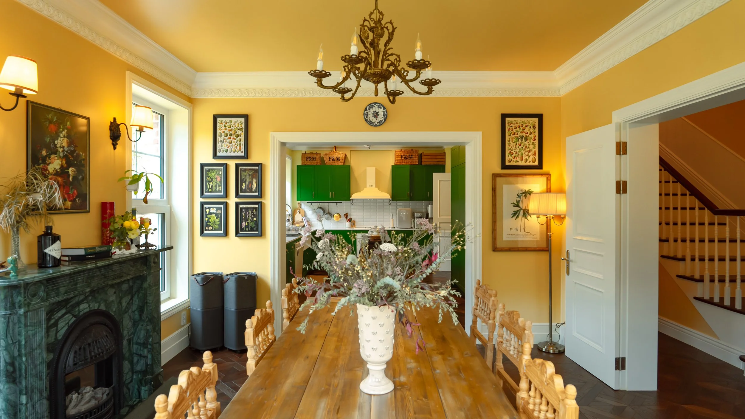 A dining room with a wooden table, floral centerpiece, yellow walls, and a view into a kitchen with green cabinets. Decor includes framed artwork, a chandelier, wall-mounted lamps, and a standing lamp.