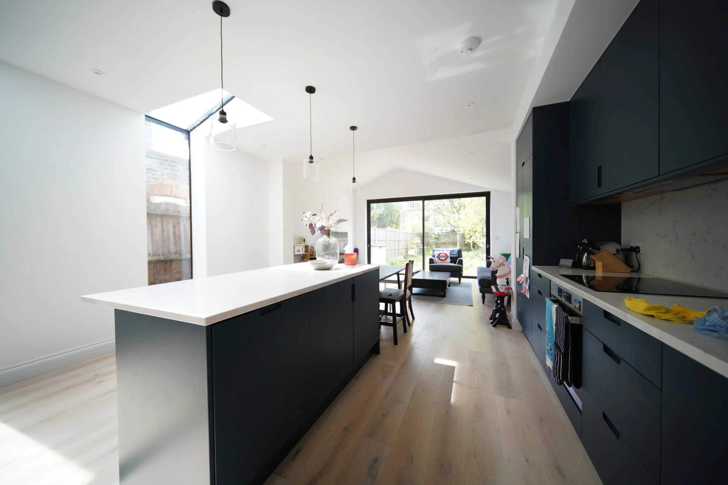 Modern kitchen with navy blue cabinets, white countertops, wooden flooring, and large windows providing natural light, with a dining table and a living area visible in the background.
