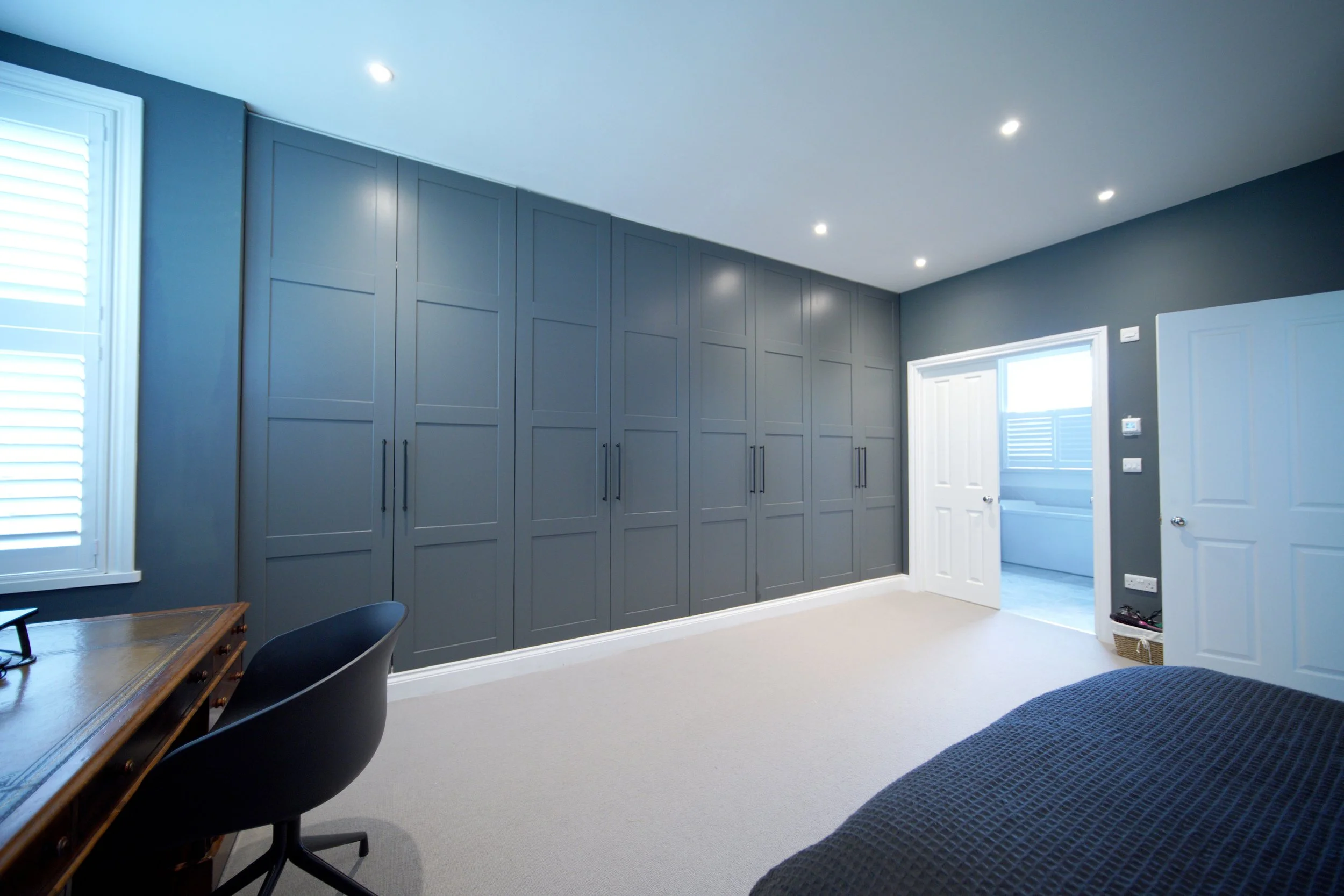 A modern bedroom with navy blue walls, a large built-in wardrobe, a window with white shutters, a desk with a chair, and a partially visible bed with a dark quilt.