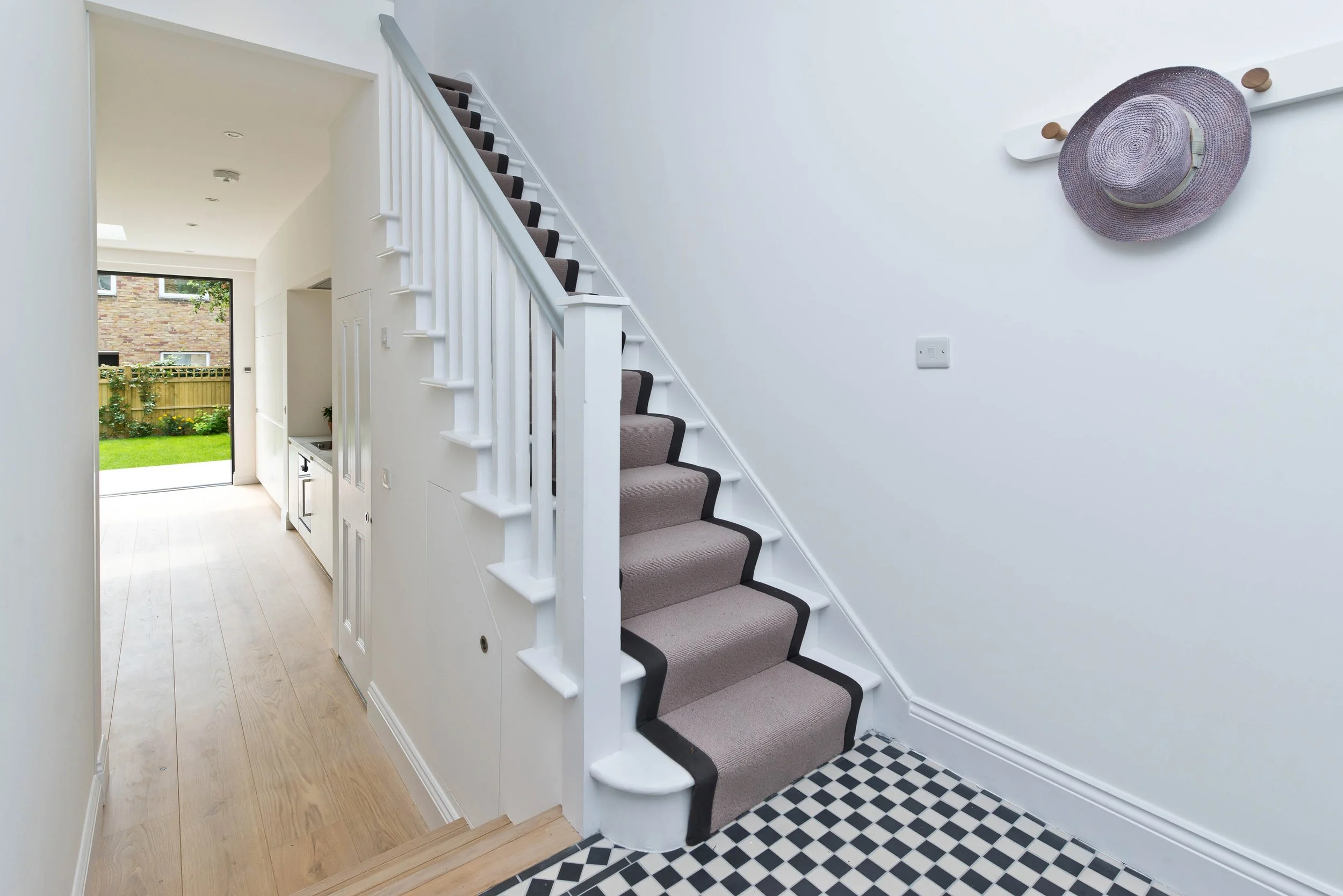 Interior view of a home entryway with a staircase, a small kitchen area, a door to the garden, and a wall with a hat on a hook.
