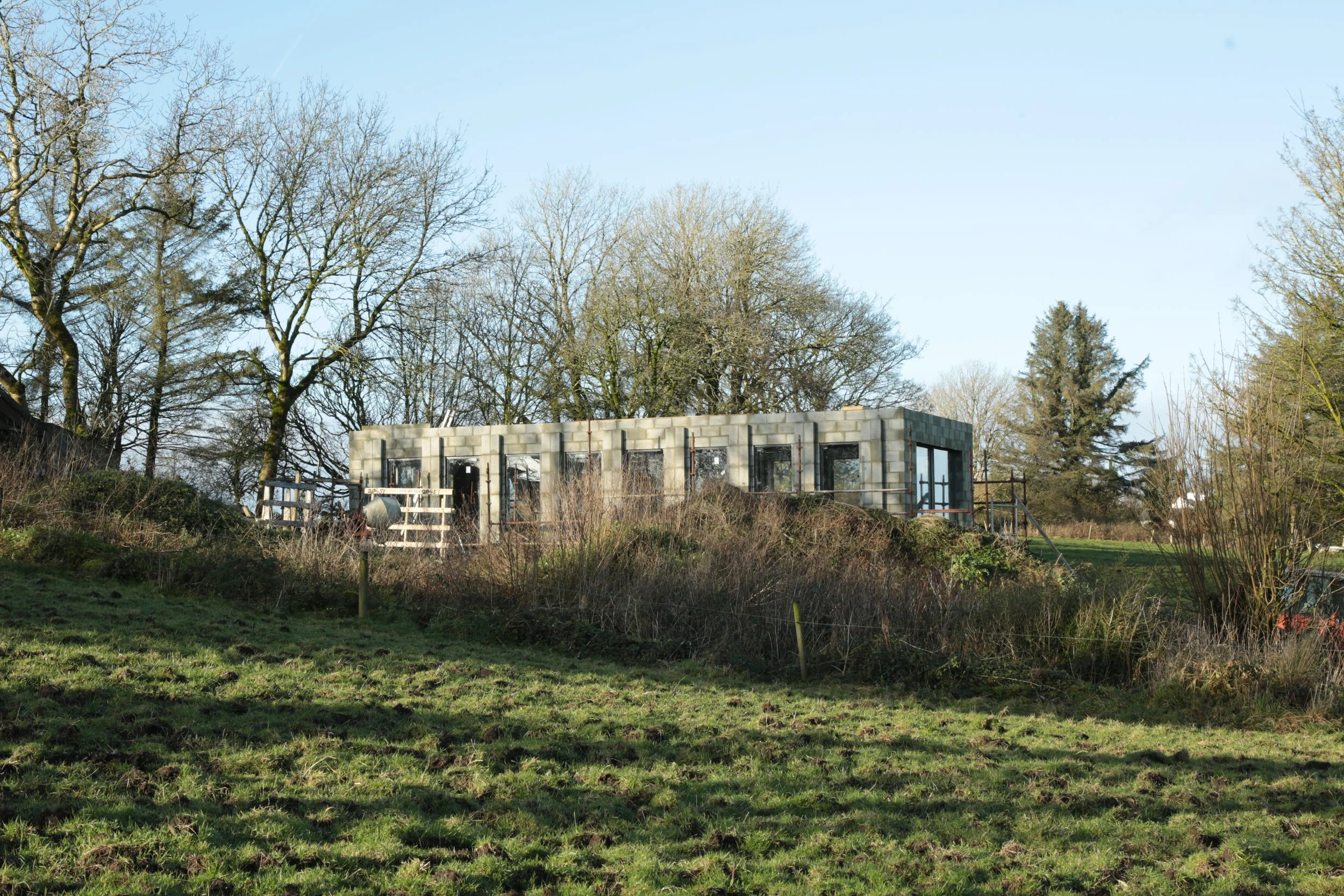 A partially constructed building on a grassy hill with trees in the background under a clear blue sky.