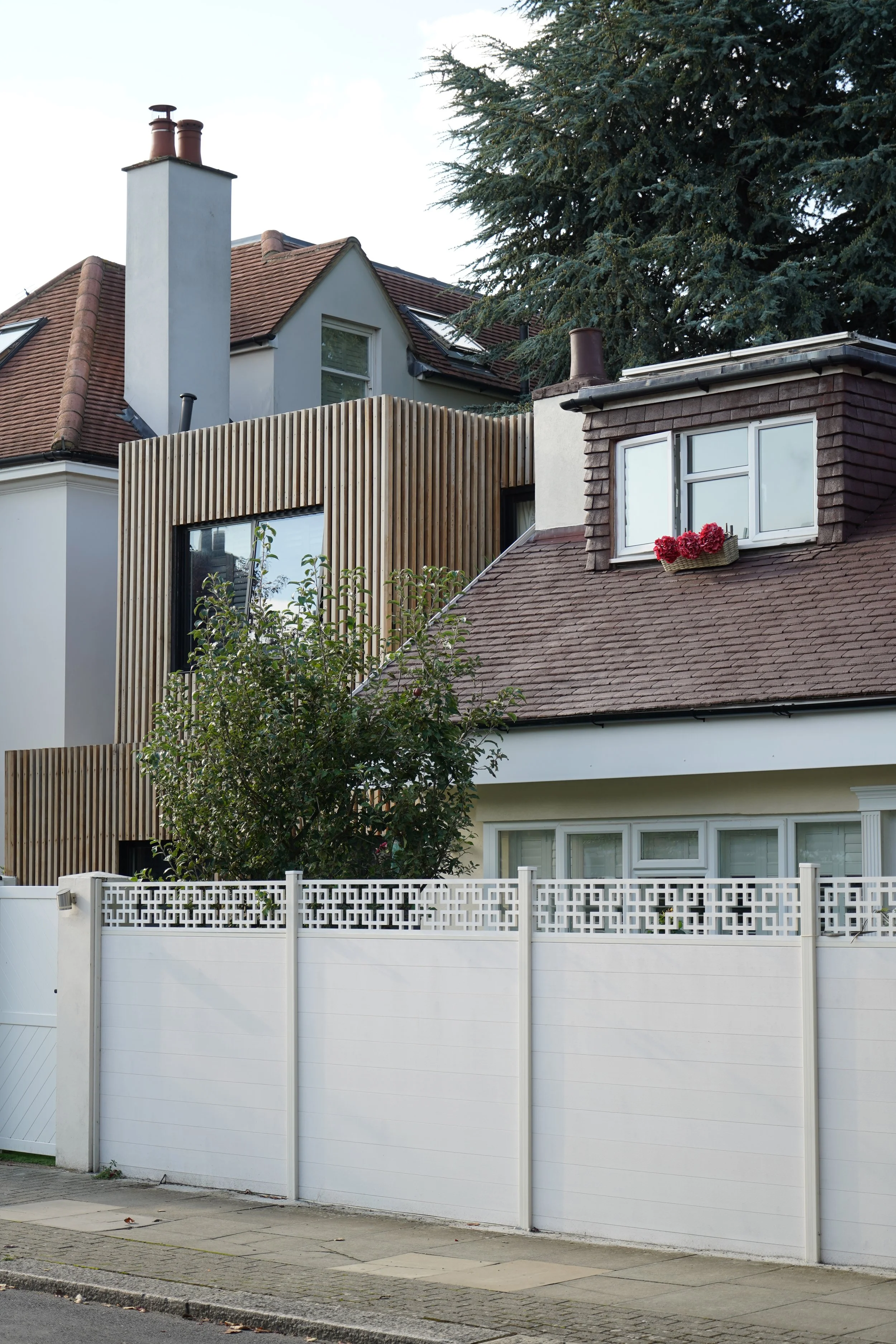 A residential scene featuring multiple houses with different architectural styles, a white fence, a sidewalk, a tree, and some flowers in a window box.