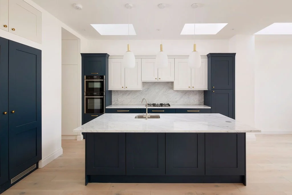 Modern kitchen with navy blue cabinets, white upper cabinets, marble countertops, and a central island. There are three pendant lights hanging above the island, and built-in appliances including double ovens.