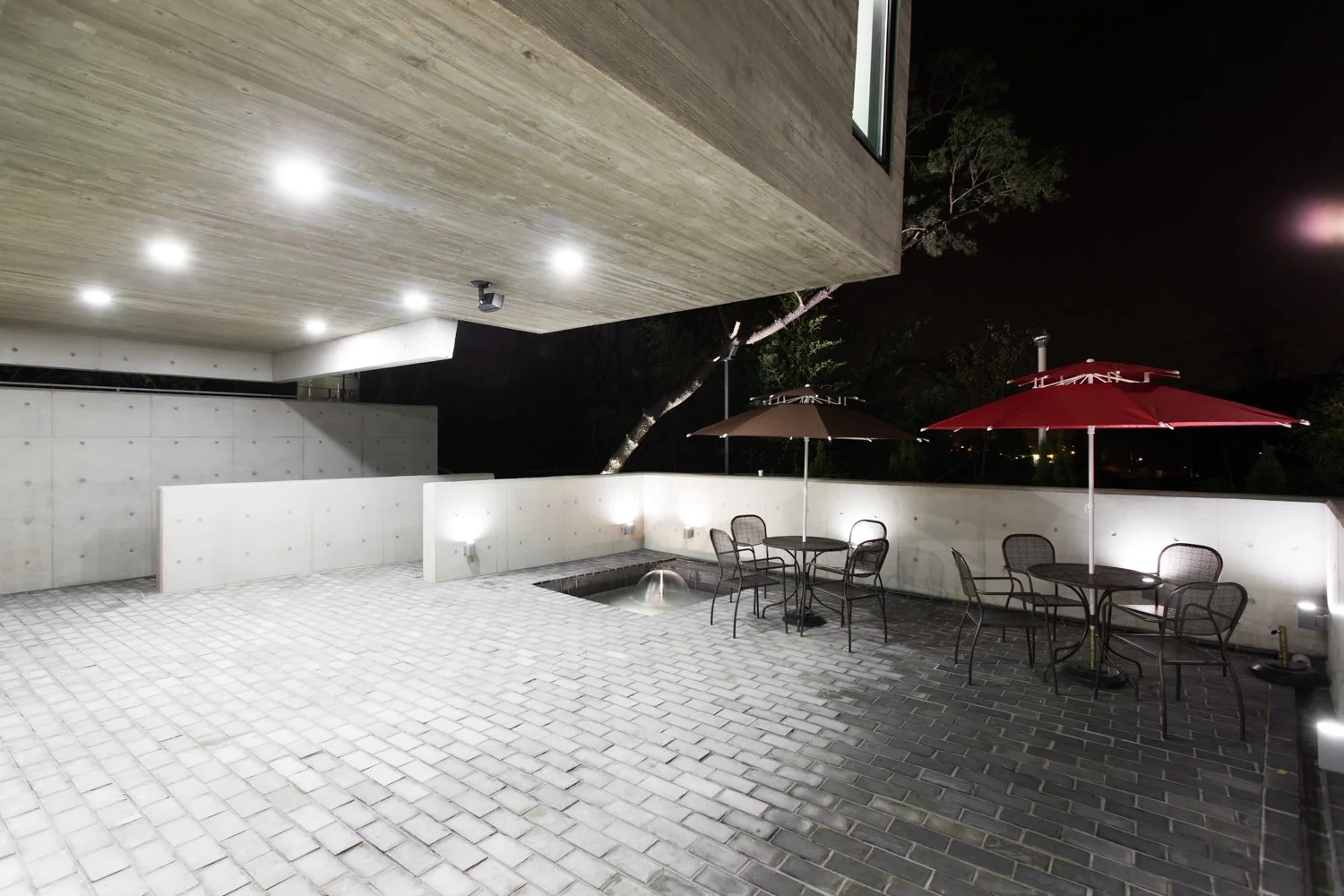 Nighttime patio with concrete and brick flooring, outdoor tables with chairs and red and beige umbrellas, illuminated lighting, modern concrete building with wood-paneled ceiling, trees in background.
