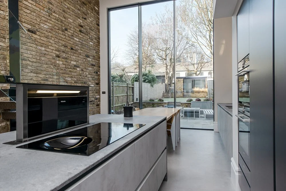 Modern kitchen with a brick accent wall, large floor-to-ceiling glass doors overlooking a backyard, minimalist cabinetry, and a black induction cooktop on a stone countertop.