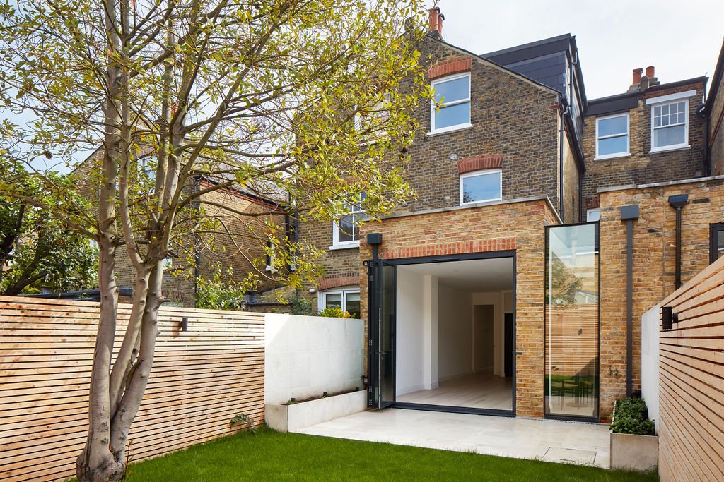 Front view of a modern brick house with a small backyard, featuring a wooden fence, a tree, green grass, large glass doors, and an outdoor patio area.