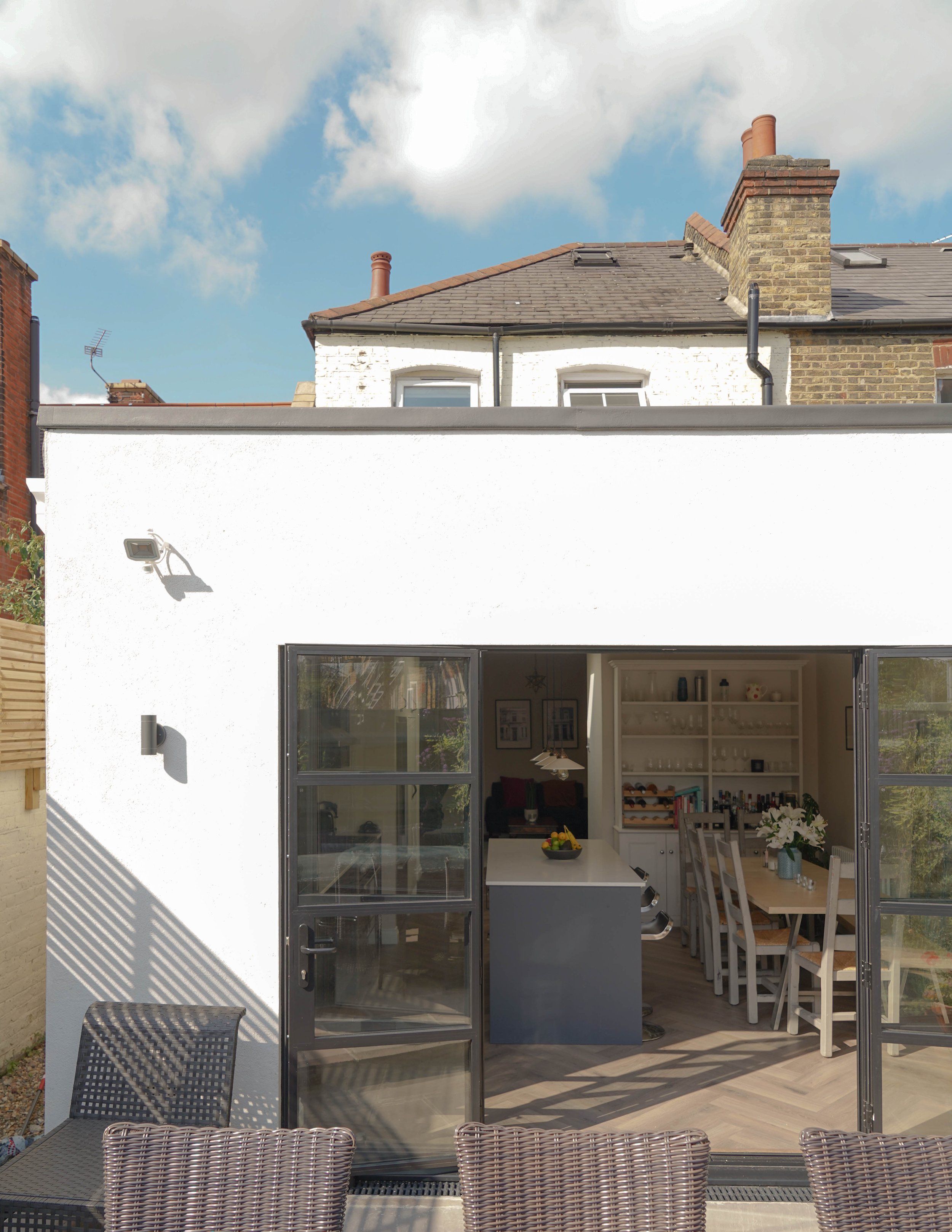 View of a white house with an open indoor dining area visible through large glass doors, outdoor patio furniture in the foreground, and a partly cloudy sky overhead.