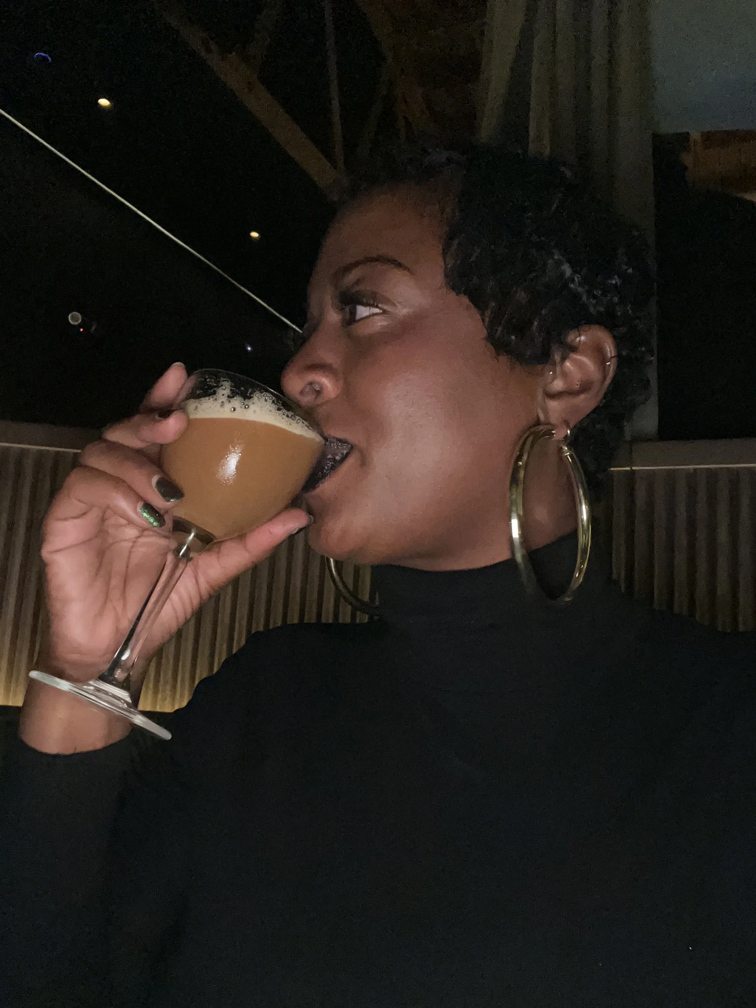 Founder of Gathered Well, Celia, side profile while wearing black turtle neck and large gold hoops. She is drinking an espresso martini.
