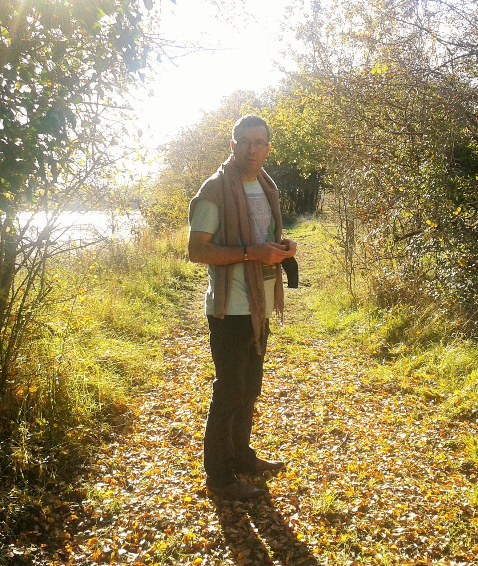 Homme marchant sur un sentier en forêt sous un soleil brillant, portant un t-shirt gris, un pantalon noir, et une écharpe ou une veste enroulée autour du cou.