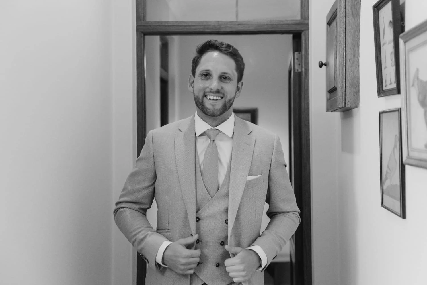 A man in a light-colored suit and tie, smiling as he adjusts his suit jacket, standing in a doorway.