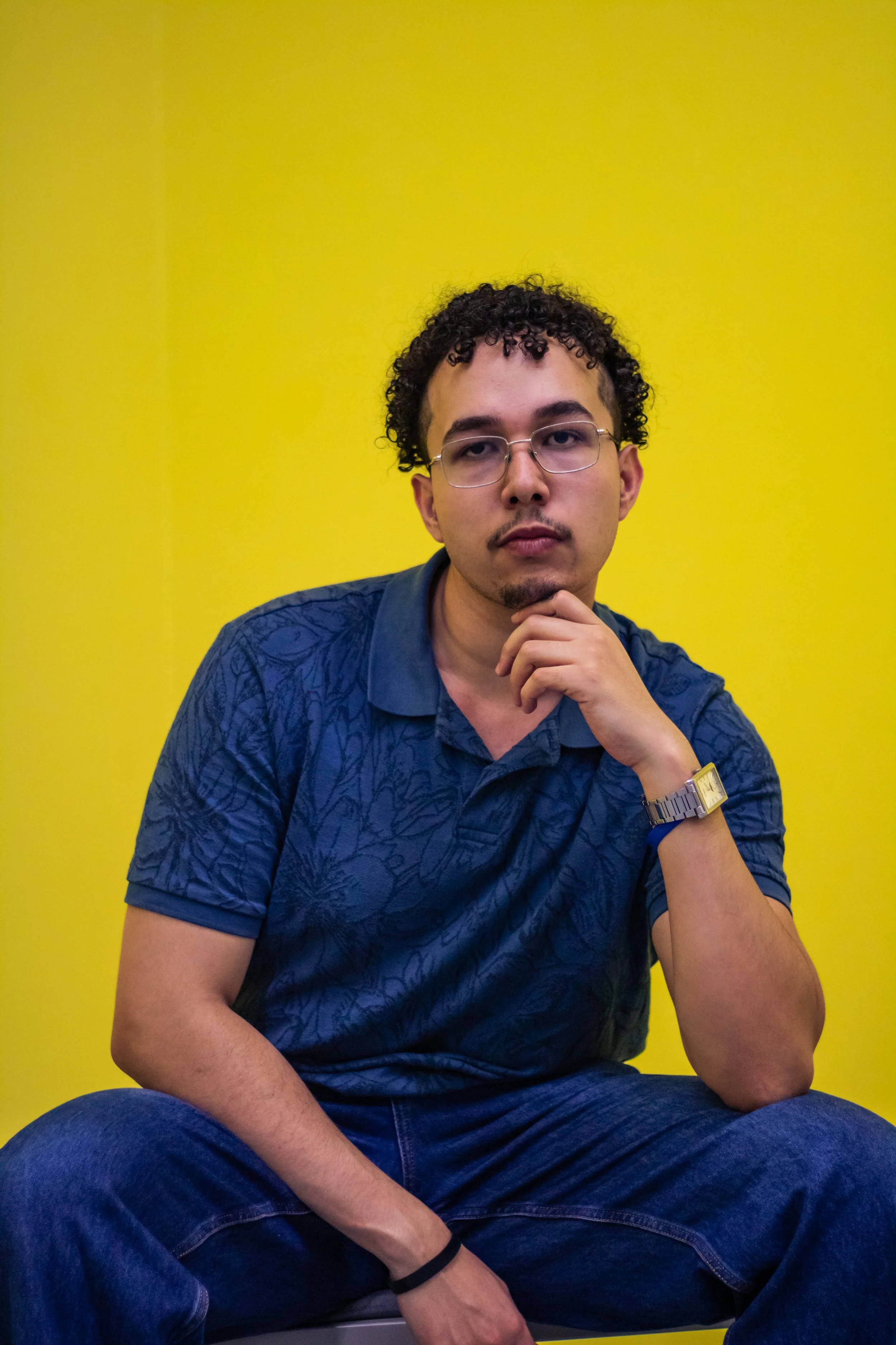 Portrait of a young man with curly hair, wearing glasses, a dark patterned shirt, and a gold wristwatch, sitting against a bright yellow wall.