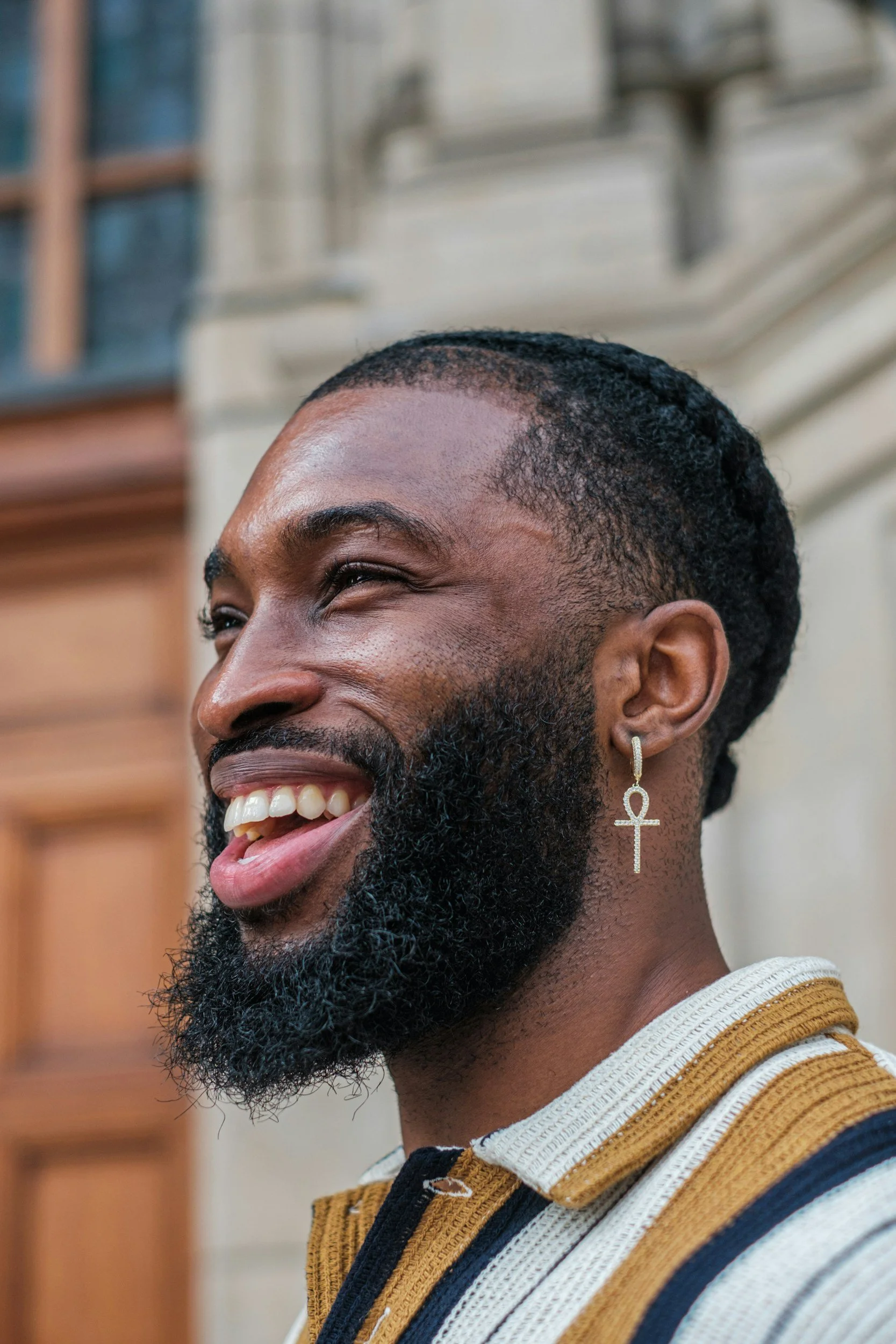 A smiling Black man with a beard, wearing an earring in the shape of an ankh and a striped shirt, standing outdoors in front of a building with a wooden door.