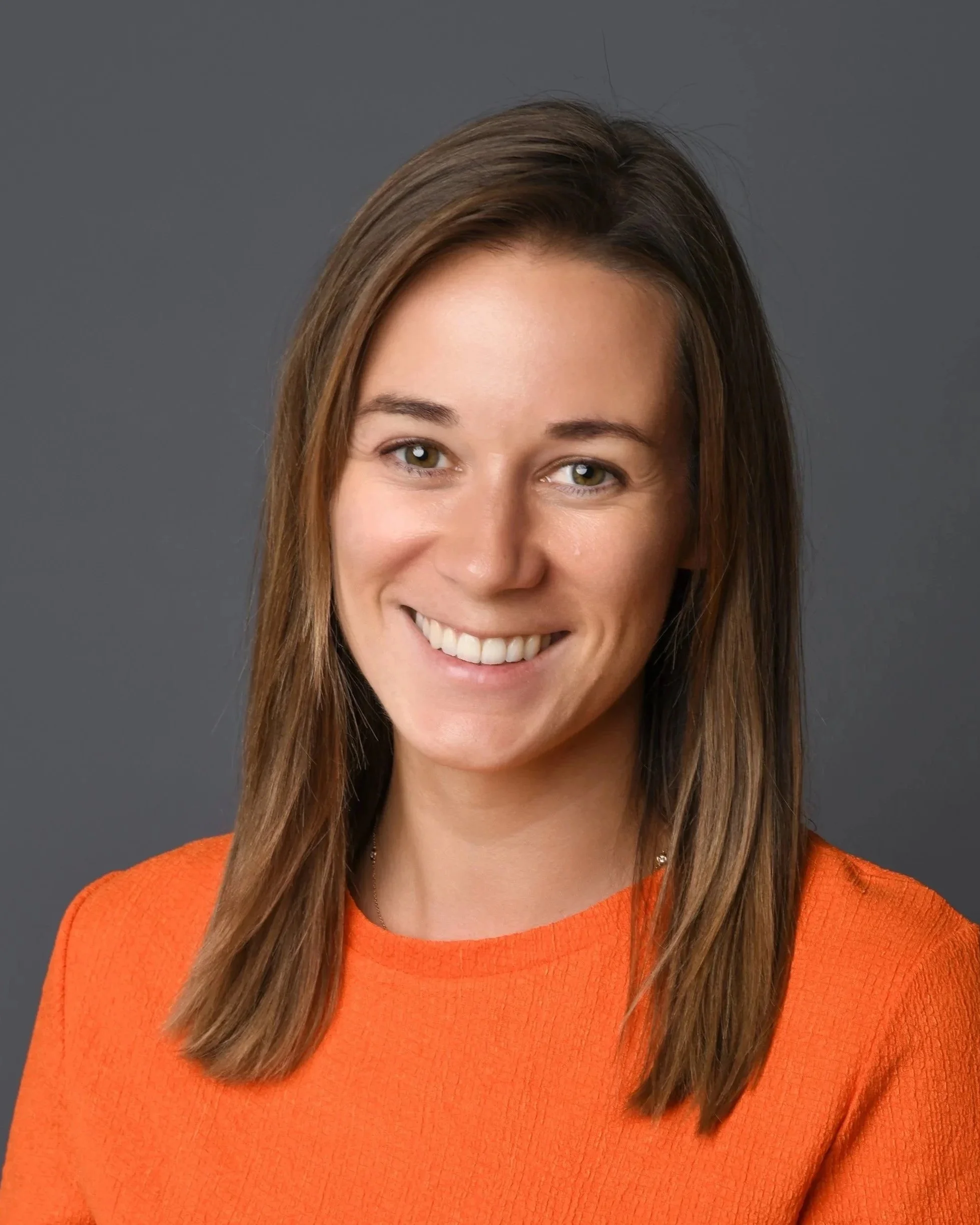A woman with shoulder-length brown hair, smiling, wearing an orange top, against a gray background.