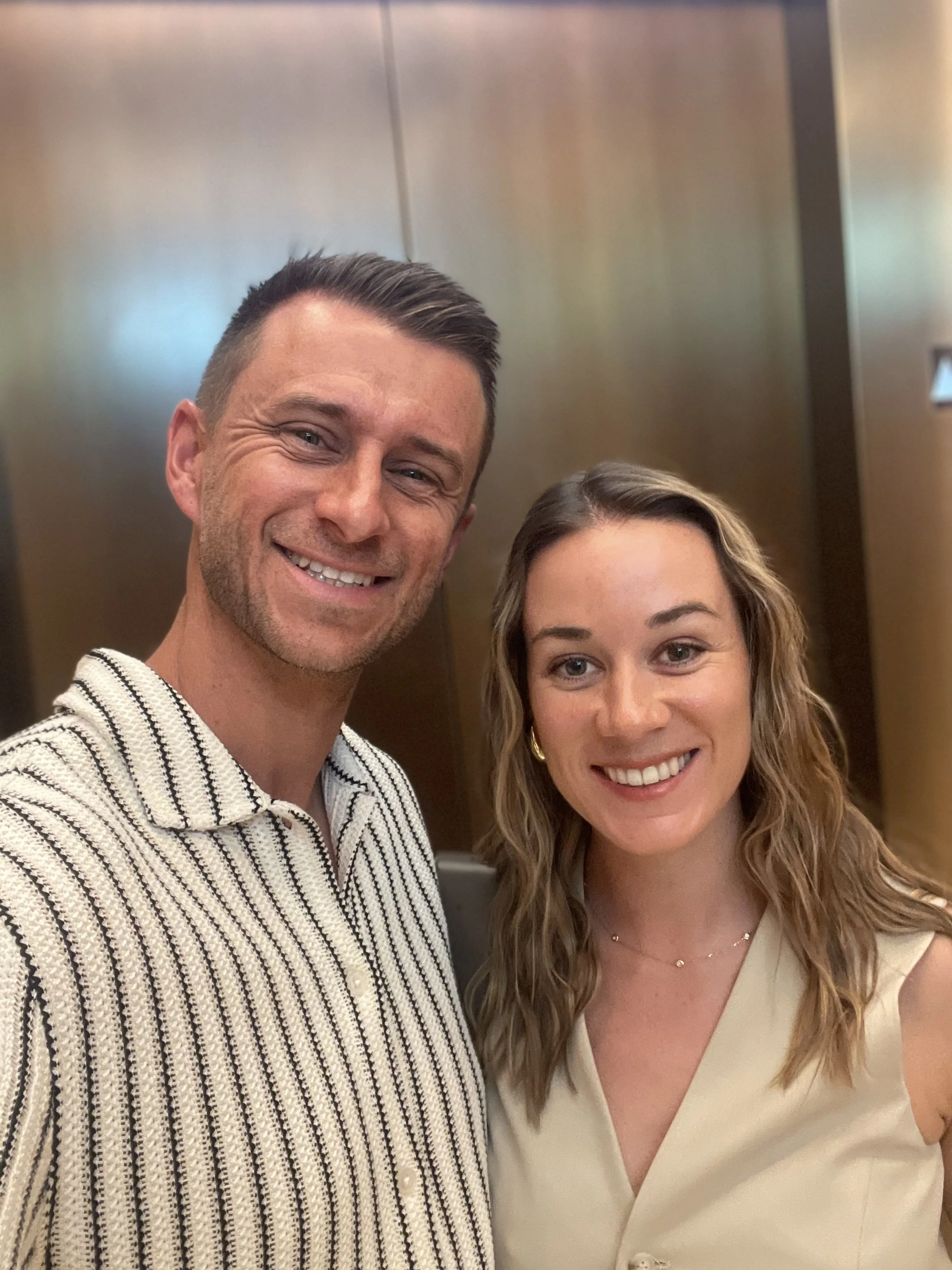 A man and woman smiling for a selfie, standing inside an elevator with metallic walls.