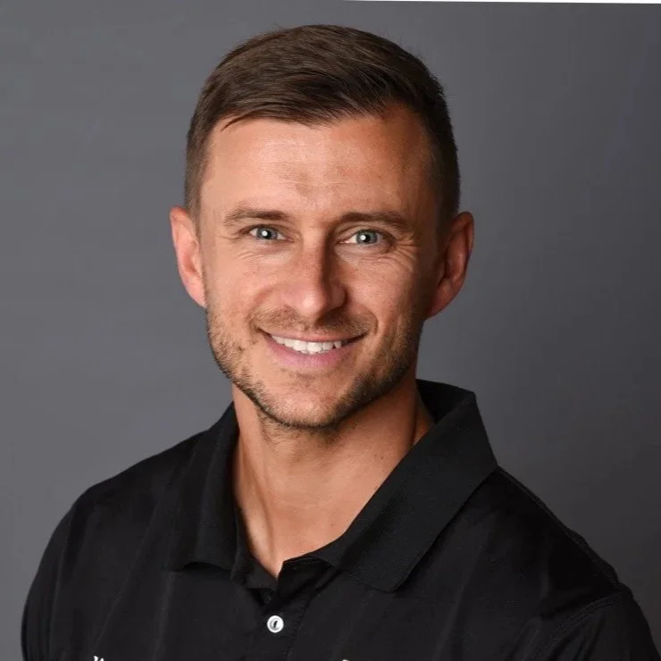 A smiling man with short brown hair, blue eyes, and a light beard, wearing a black polo shirt, against a gray background.