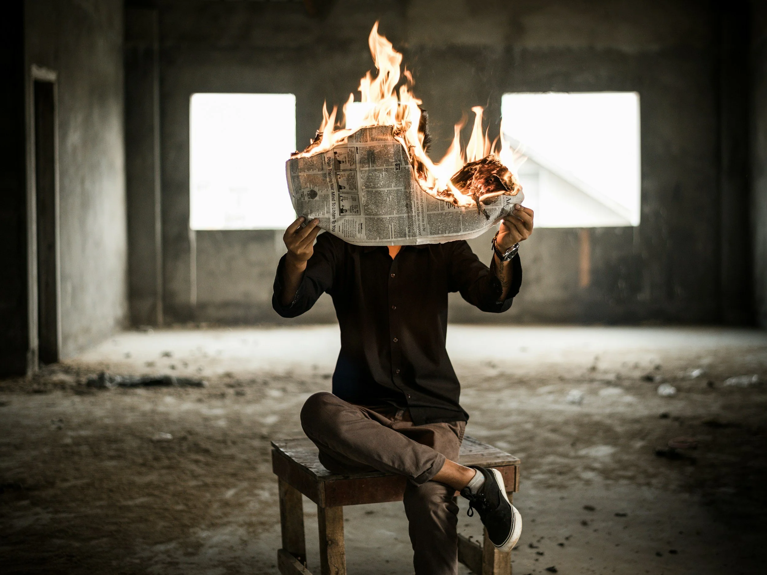 Person sitting cross-legged on a wooden stool in an unfinished room, holding a burning newspaper with flames, dark clothing, and large windows in the background.