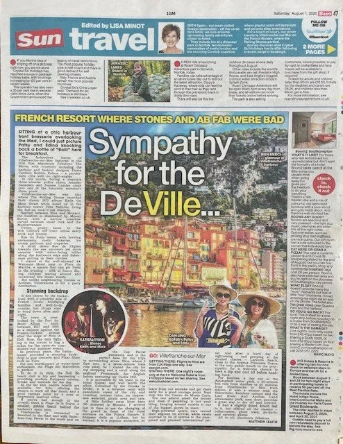 A newspaper travel section featuring an article titled 'Sympathy for the DeVille.' The headline is in large white and yellow letters on a blue background. The page includes a color photo of two women in sunglasses and hats, smiling near a waterfront with colorful buildings in the background. The article discusses a French resort with a backdrop of colorful buildings and mentions the Villefranche-sur-Mer.