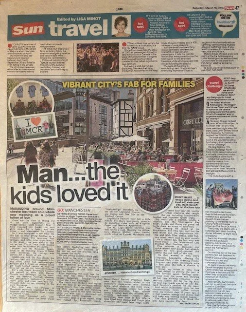 A newspaper page featuring a headline about Manchester with a large image of a lively city street with outdoor seating, shops, and people walking. Smaller images include a close-up of children, a building, and a photo of a colorful outdoor scene.