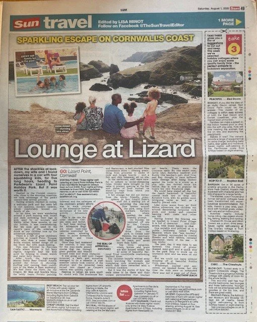 A newspaper page from the Sun travel section, featuring a main story titled 'Lounge at Lizard.' The page includes a large photo of people sitting on a rocky beach near the ocean, with rocks and waves in the background. There are also smaller images and columns of text discussing travel destinations like Cornwall and nearby areas.