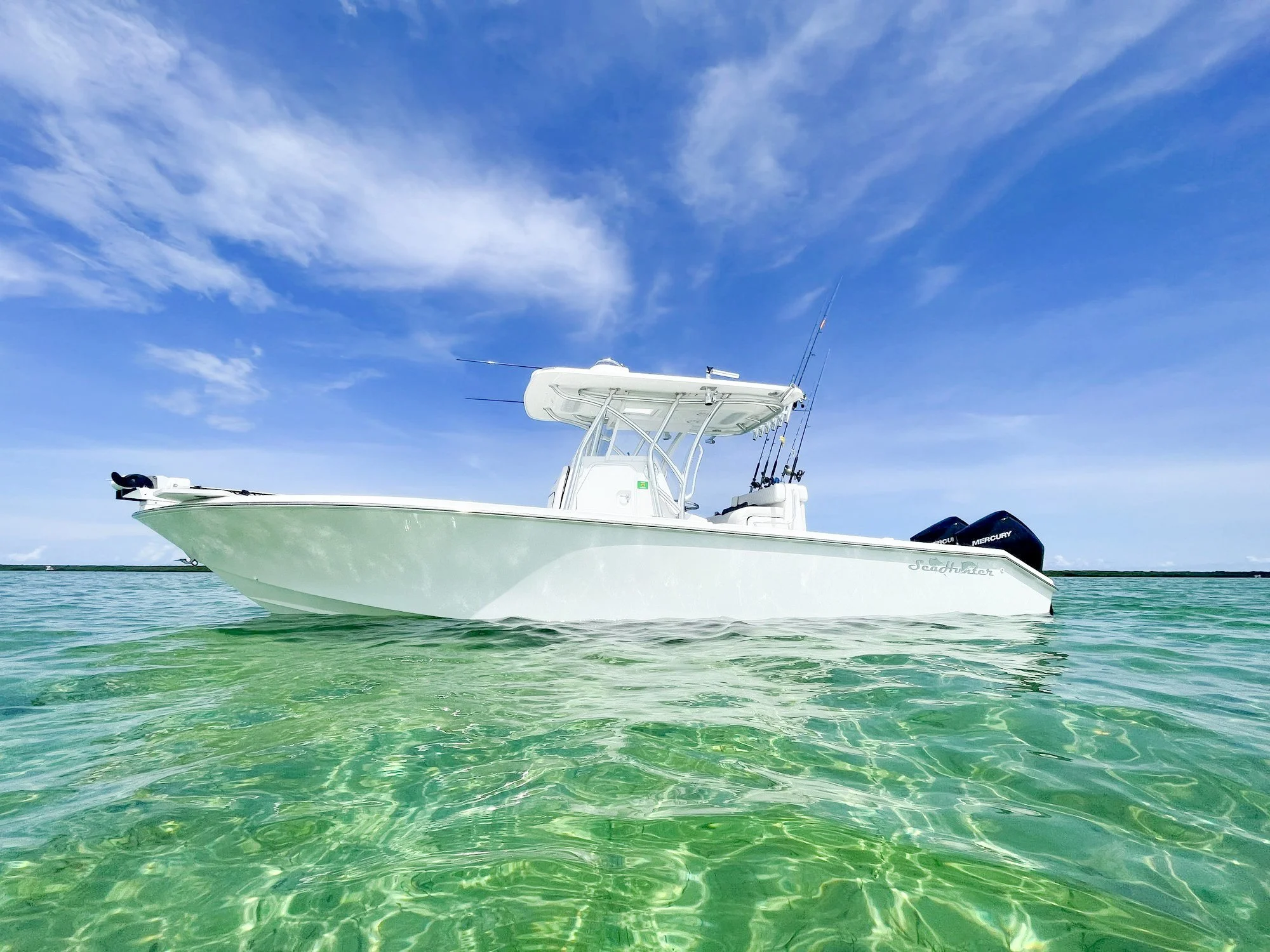 A white fishing boat with mounted fishing rods sailing in clear turquoise waters under a partly cloudy blue sky.