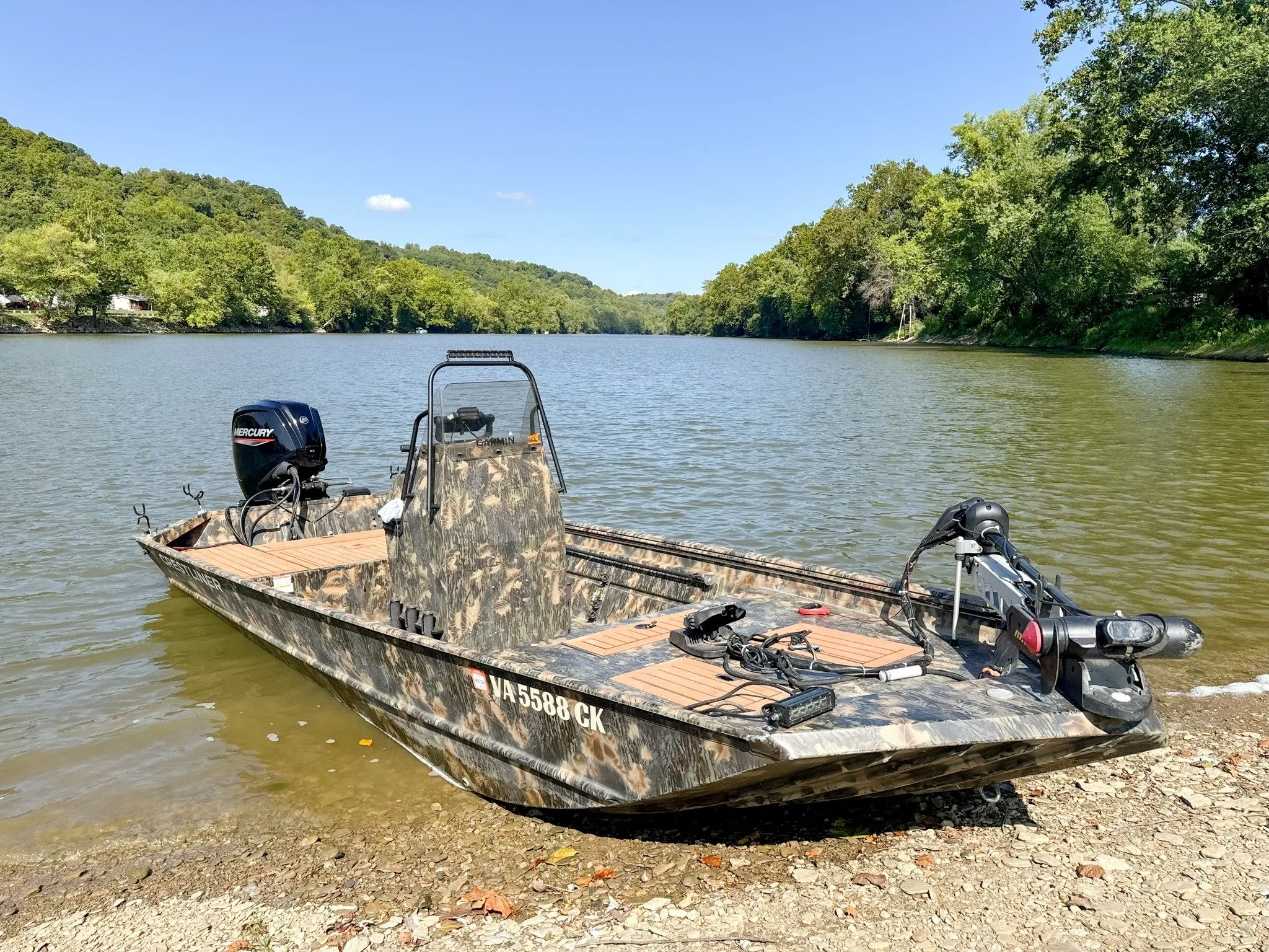 A camouflage-colored boat with a Mercury motor and a built-in windshield docked on a lake shoreline surrounded by green trees under a blue sky.