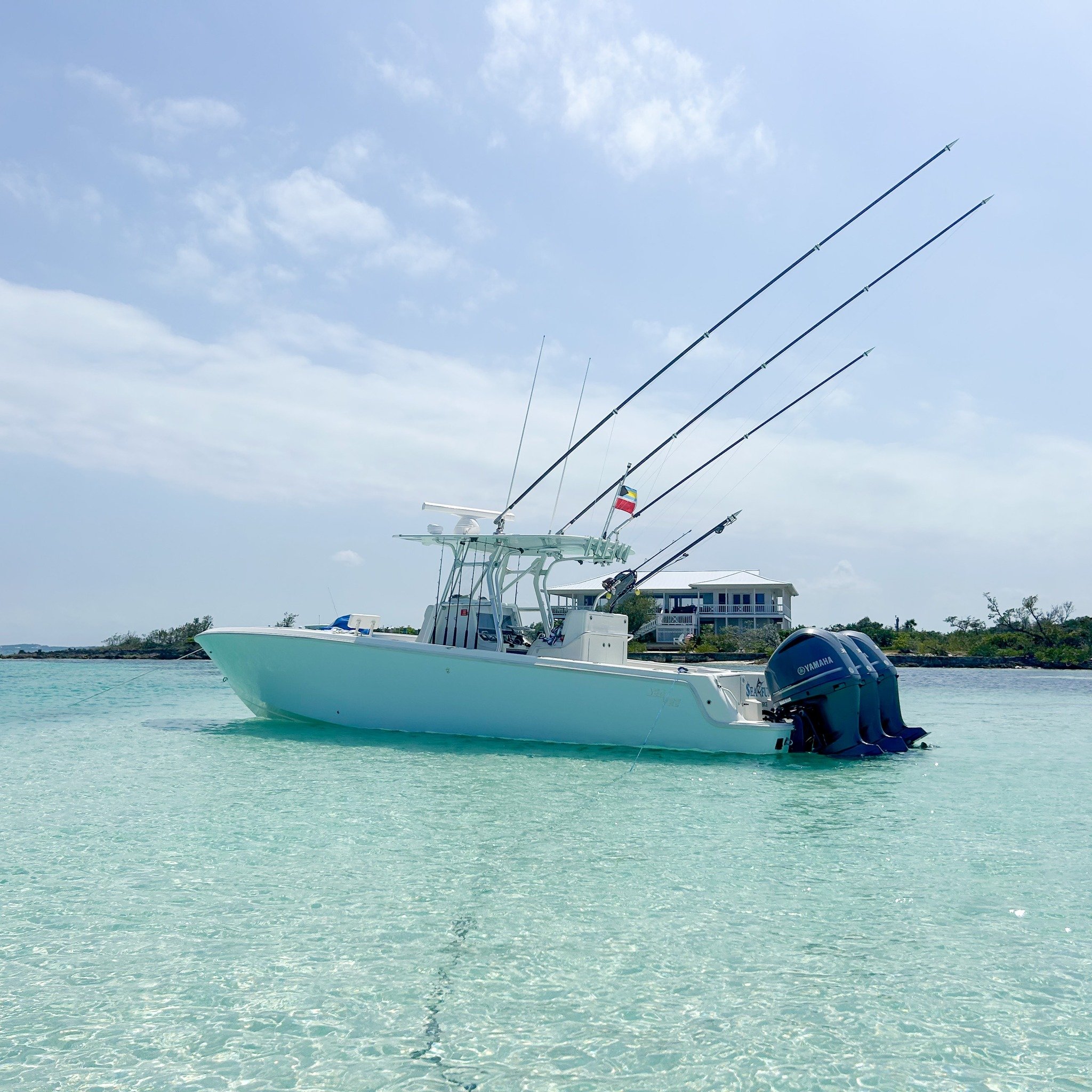 A boat with multiple fishing rods floating in clear, shallow ocean water near a shoreline.