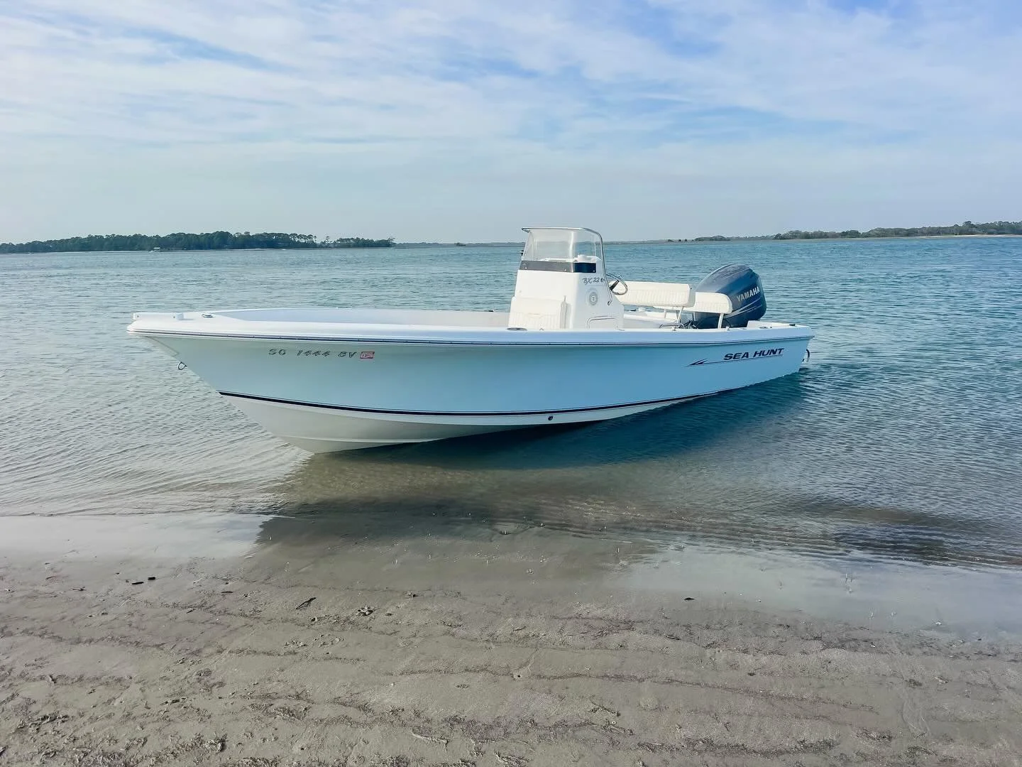 White Sea Hunt boat with Yamaha outboard motor anchored near sandy beach on calm water with distant shoreline and partly cloudy sky.