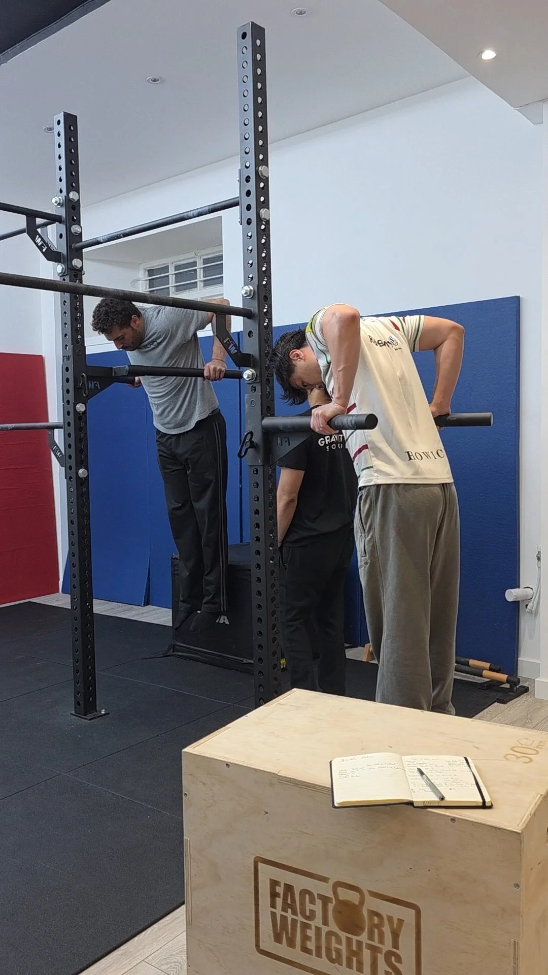 Two men are using a power rack for exercise; one is on a box doing dips, the other is holding onto the rack for support, and a third person is behind them. A notebook and pen are on a wooden box labeled 'Factory Weights' in the foreground.