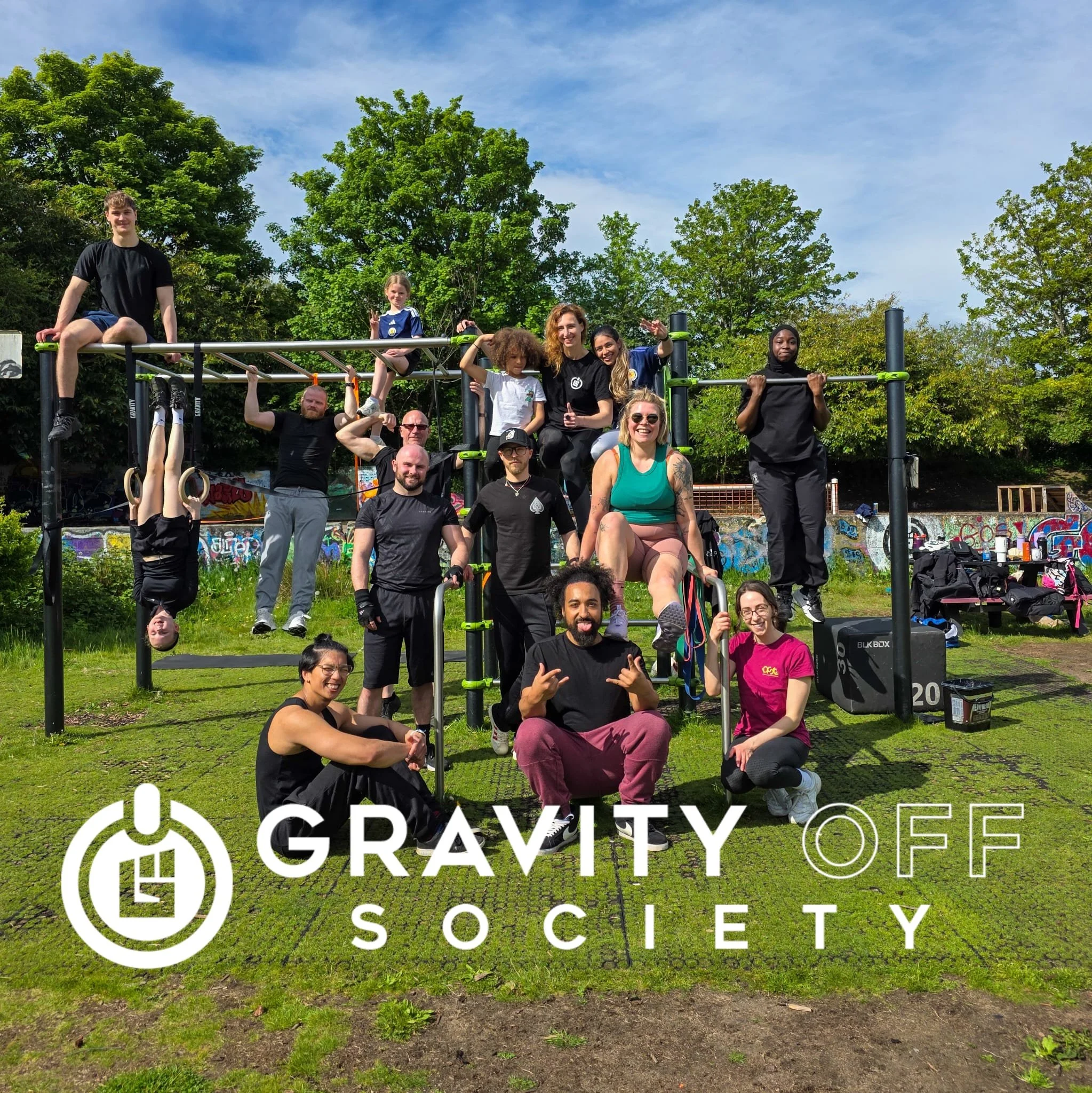 Group of diverse people, including children and adults, posing outdoors in front of a playground structure with graffiti-covered wall underneath. Some are sitting, standing, or hanging on gymnastic rings, smiling and making peace signs. Bright, sunny