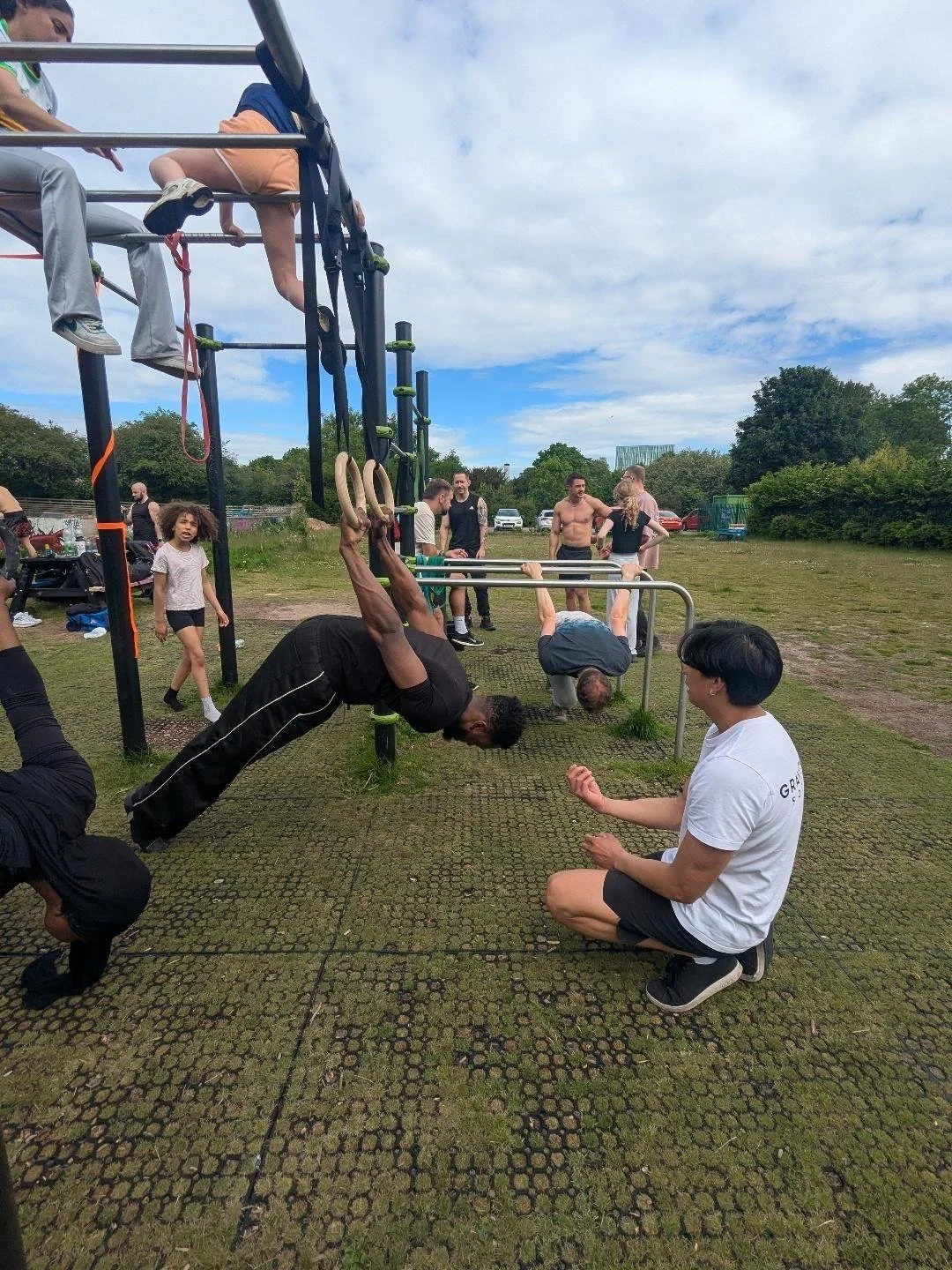 People working out on outdoor fitness equipment, including rings and bars, in a park on a cloudy day.