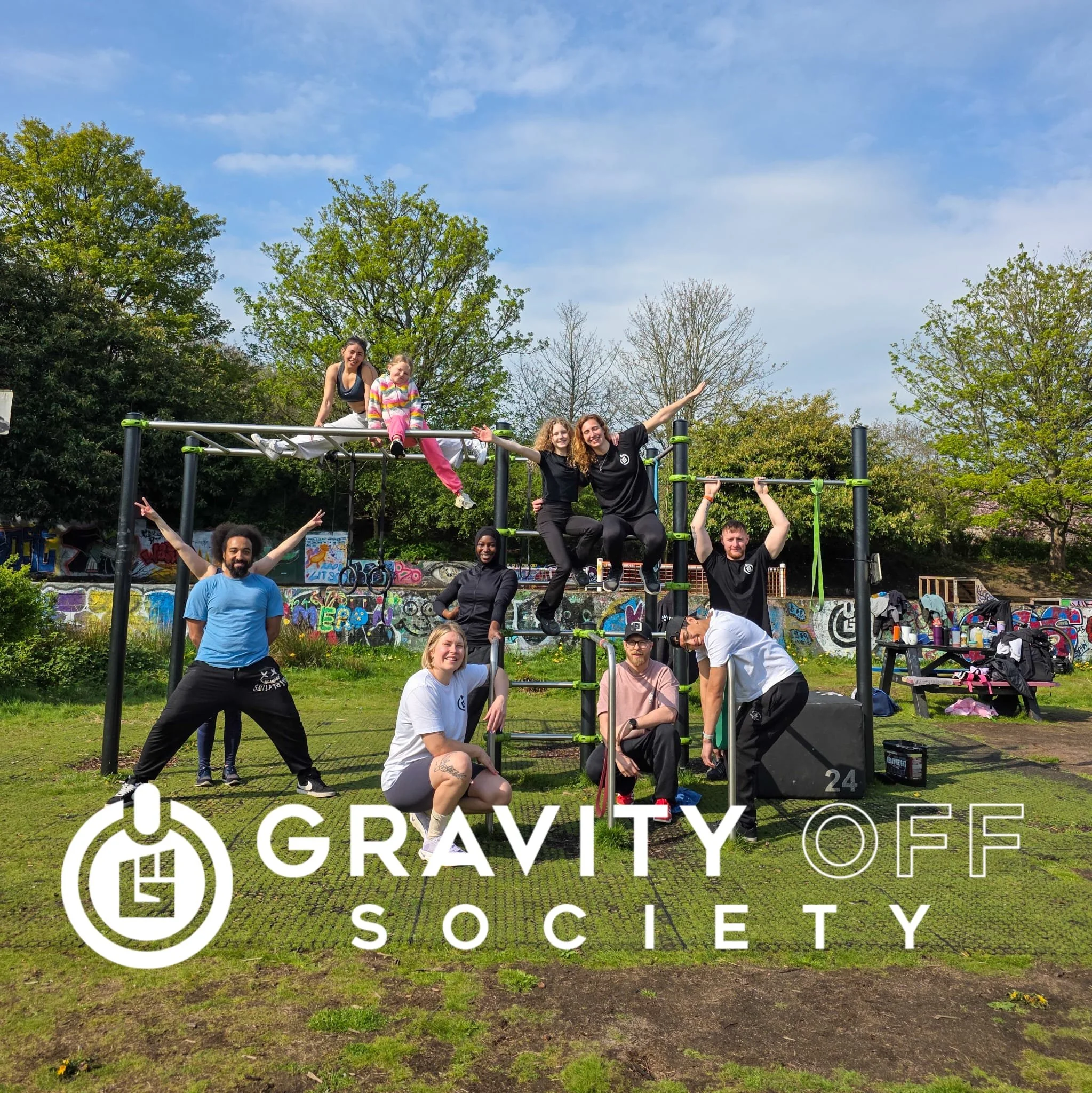 Group of people participating in a fitness event outdoors, with a large 'Gravity' Society sign and graffiti-covered wall in the background, under a blue sky with some clouds.